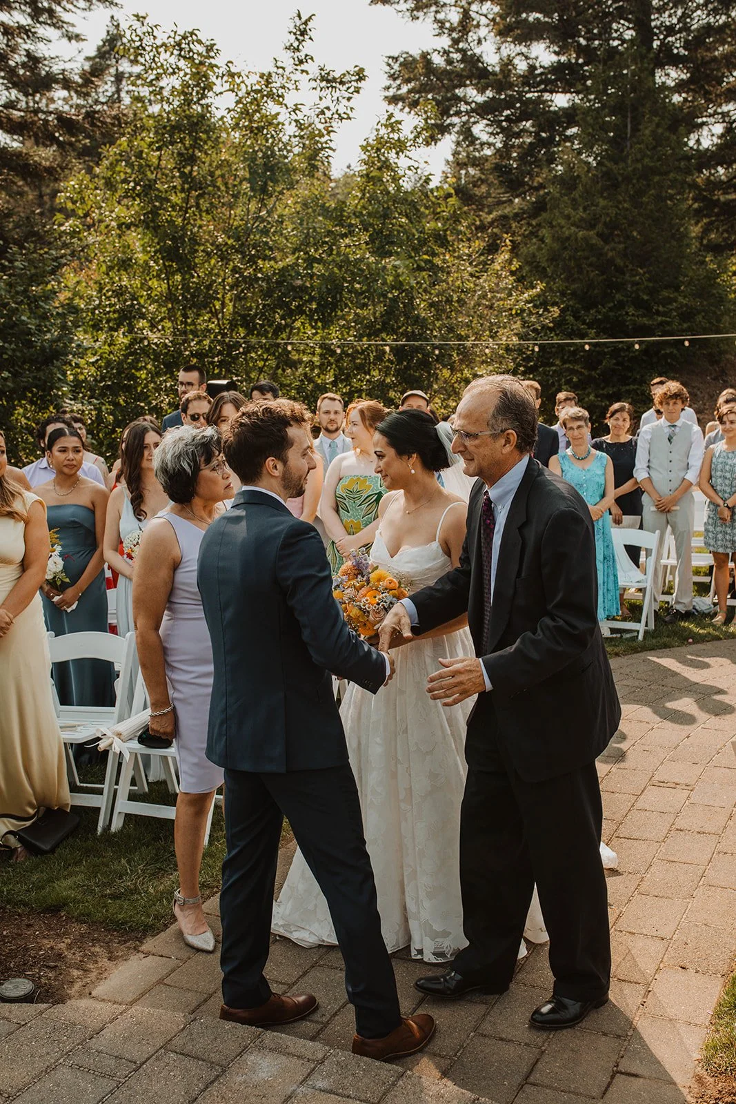Groom shakes hands with a family member while the bride smiles beside him after an outdoor wedding ceremony surrounded by guests and greenery.