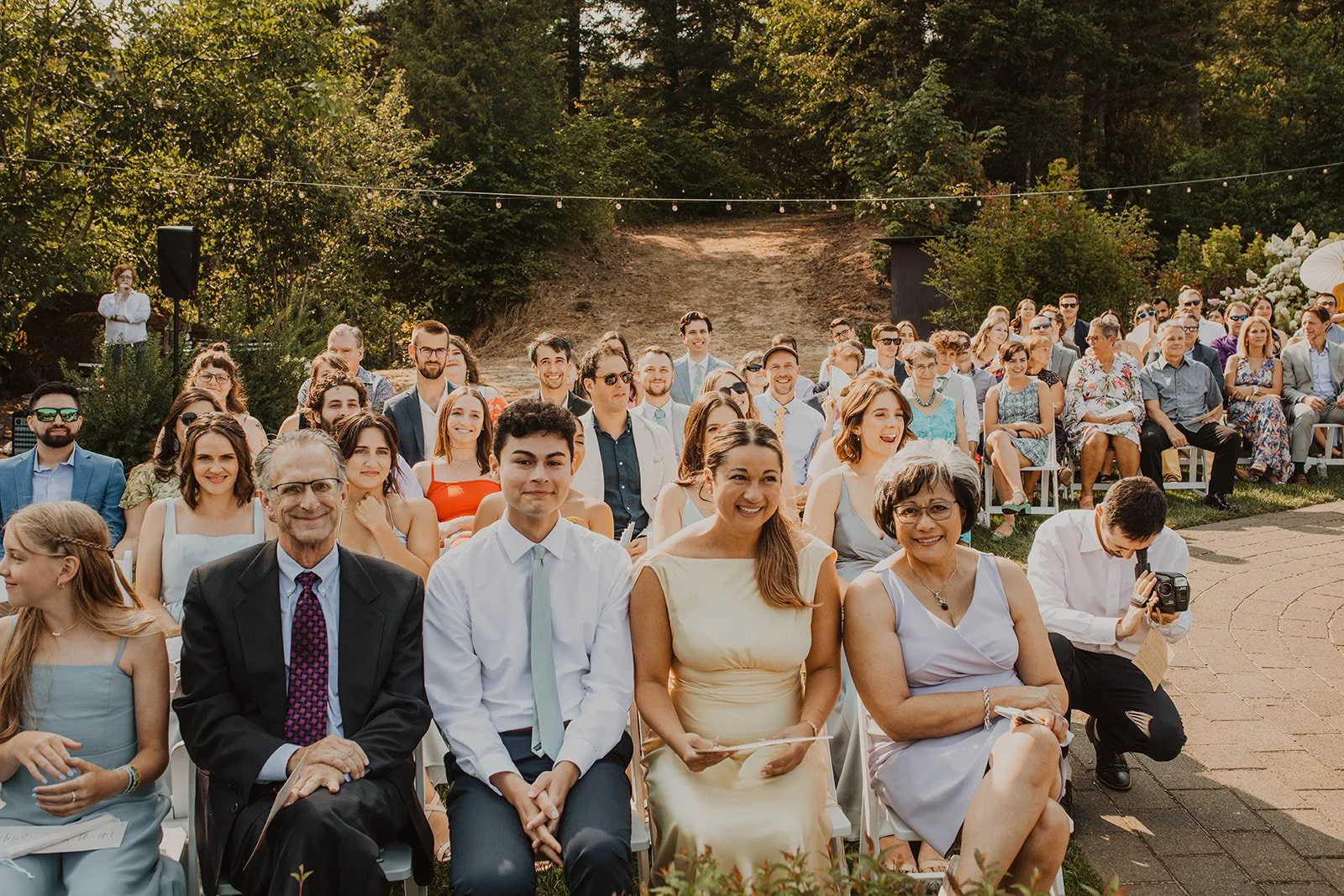 Wedding guests smile and wait for the ceremony to begin during an outdoor celebration surrounded by trees and warm afternoon light.