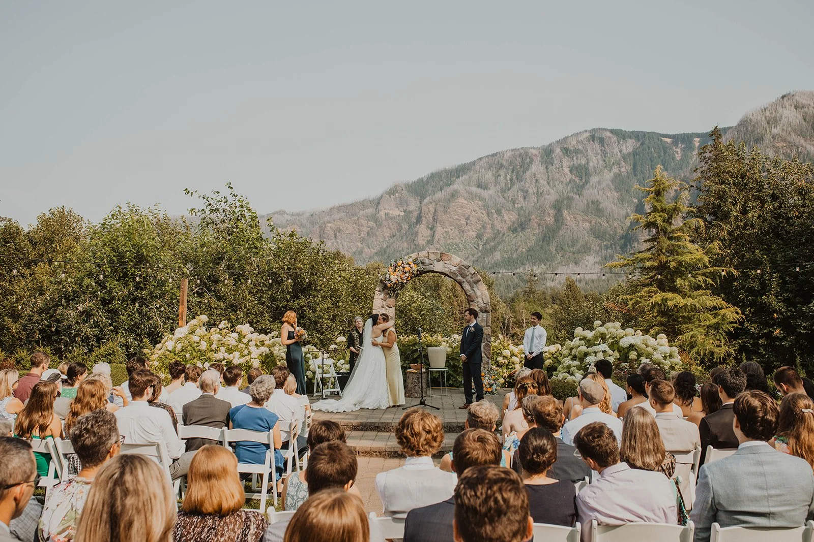 Wide shot of an outdoor mountain wedding ceremony with guests seated beneath a floral stone arch surrounded by lush greenery and scenic views.