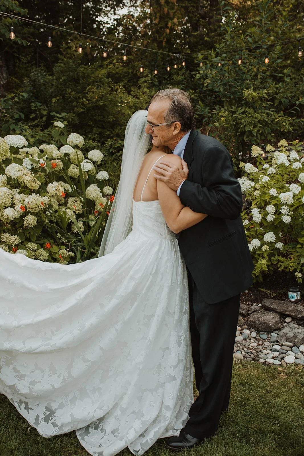 Bride shares an emotional hug with her father during a first look in a garden surrounded by white hydrangeas and string lights at Cape Horn Estate.