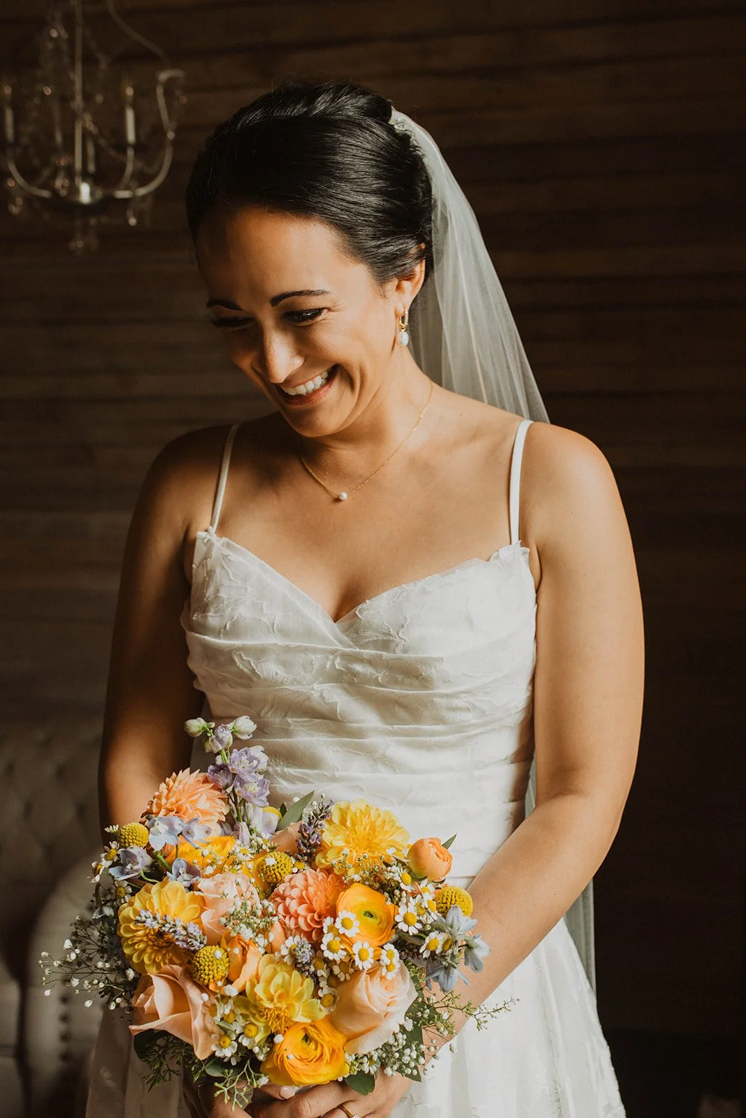 Bride smiles while holding a colorful bouquet during bridal portraits inside a rustic wood-paneled getting-ready suite.
