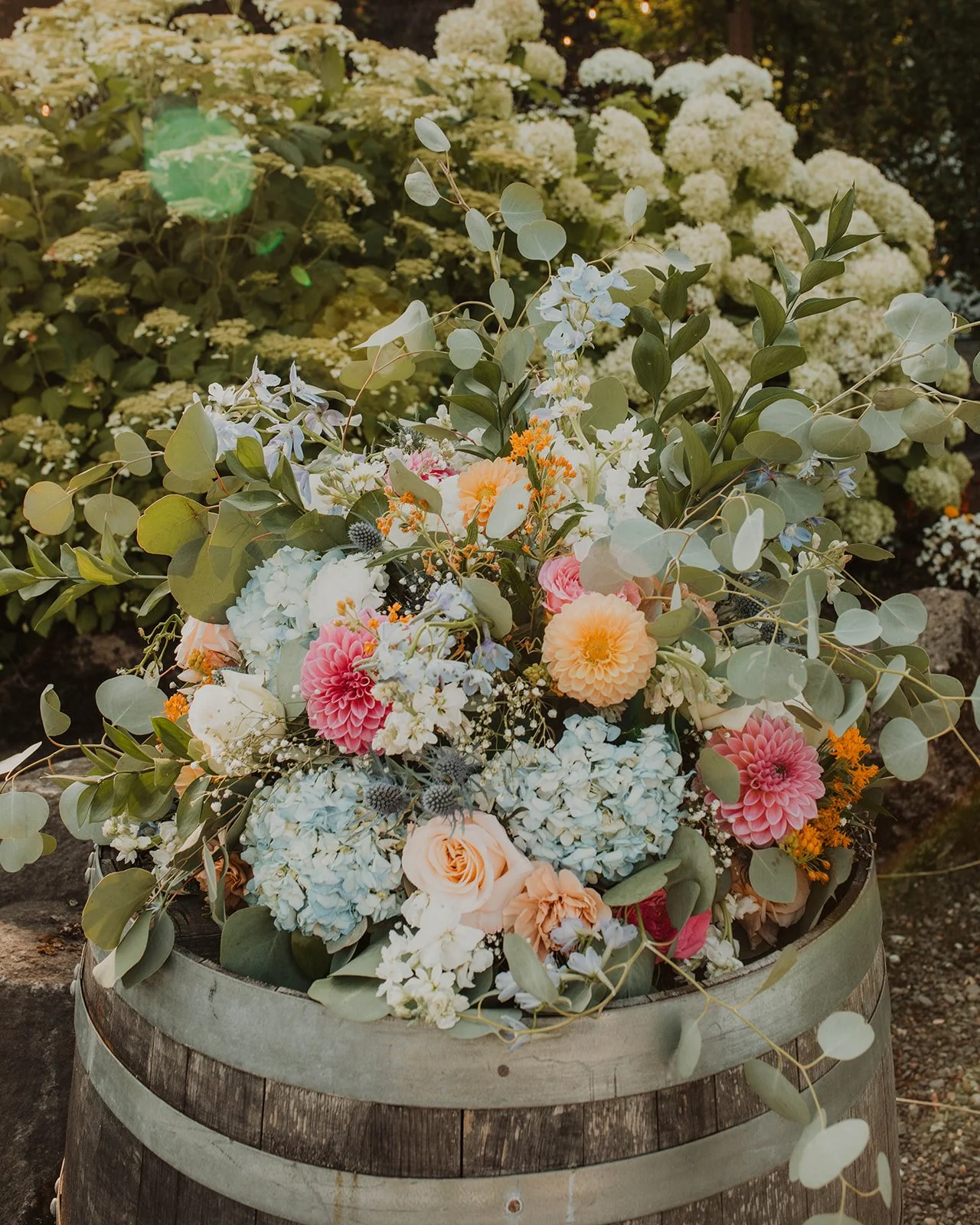 Soft pastel wedding floral arrangement displayed in a rustic wooden barrel with hydrangeas, dahlias, roses, eucalyptus, and wildflower accents at an outdoor garden wedding venue