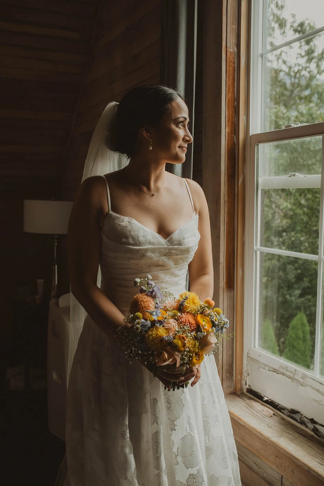 Bridal portrait of a bride holding a colorful wildflower bouquet while standing beside a softly lit window at Cape Horn Estate.