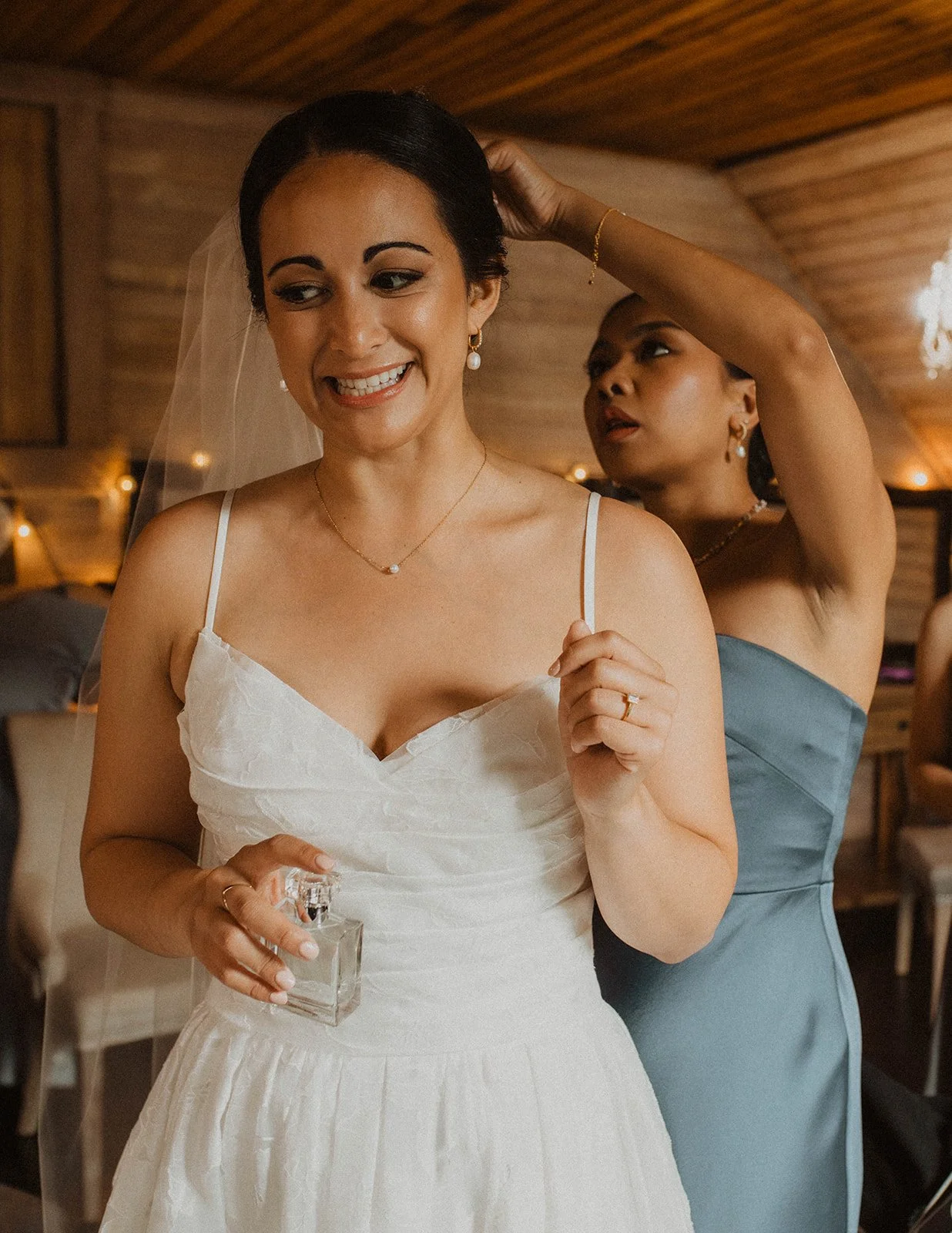 Bride smiles while a bridesmaid adjusts her veil during wedding preparations inside a cozy wood-paneled bridal suite.