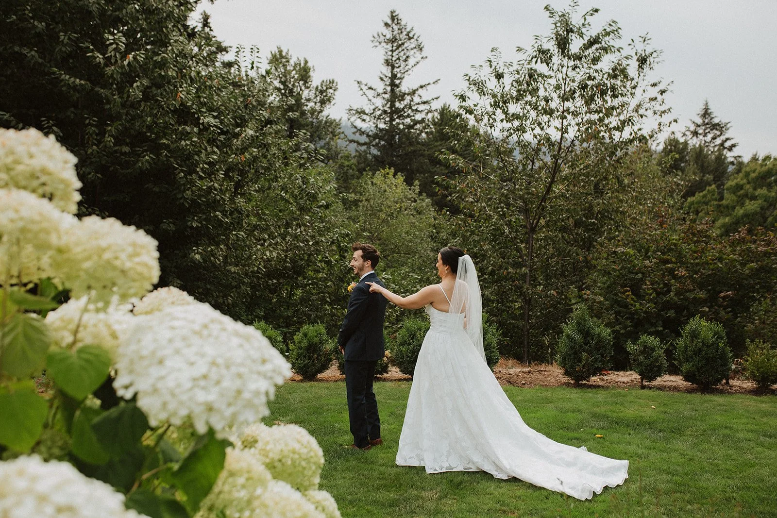 Bride in a flowing wedding gown approaches the groom from behind for their outdoor first look in a garden setting.