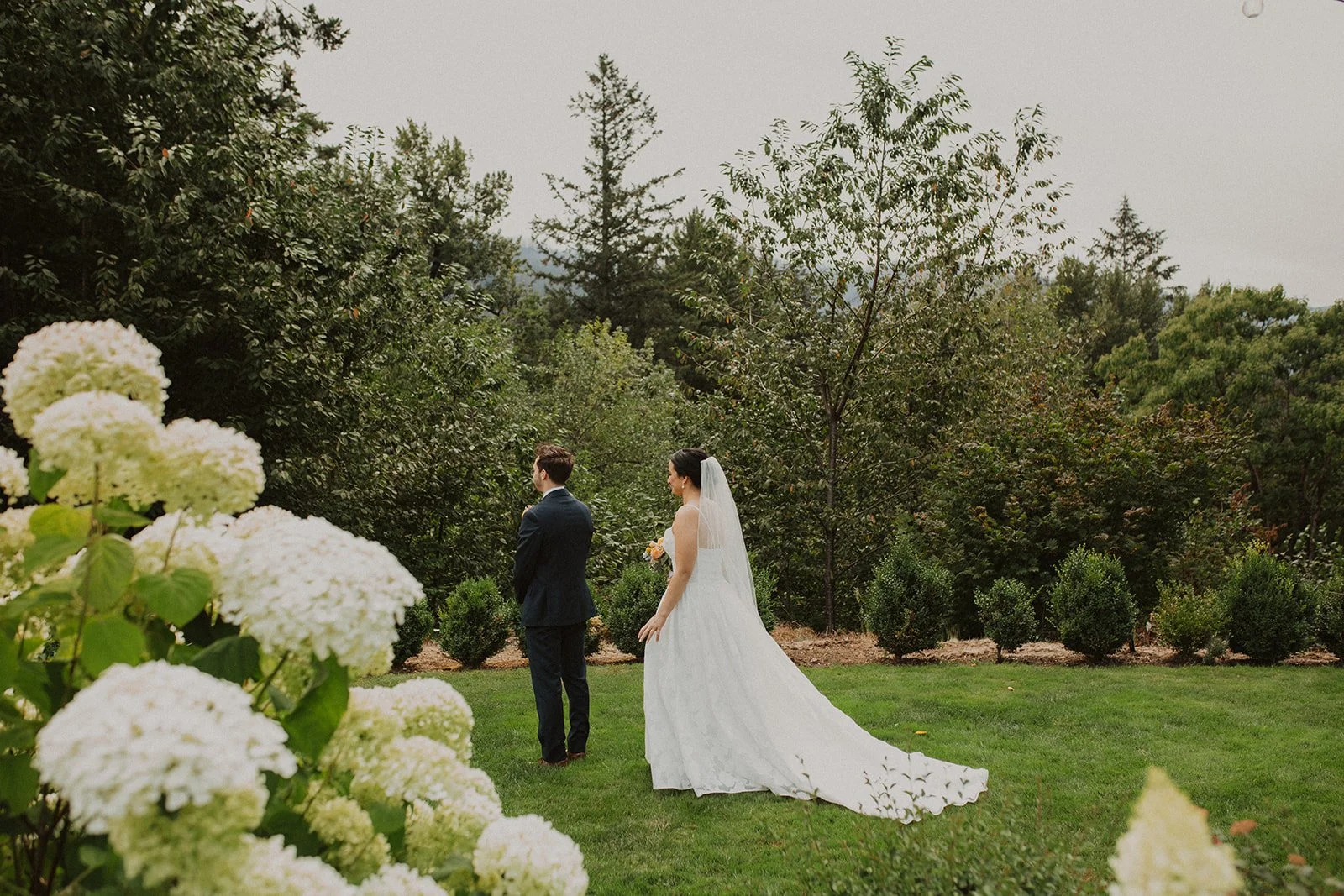 Bride taps the groom on the shoulder before their first look in a romantic outdoor wedding garden with blooming hydrangeas.