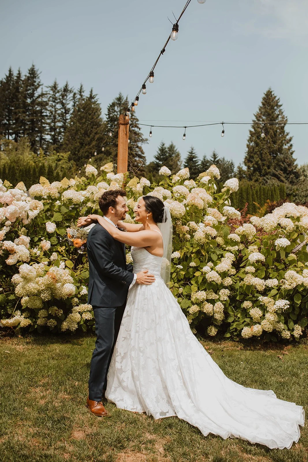 Newlywed couple embraces and smiles in front of blooming white hydrangeas and string lights during their outdoor wedding portraits.