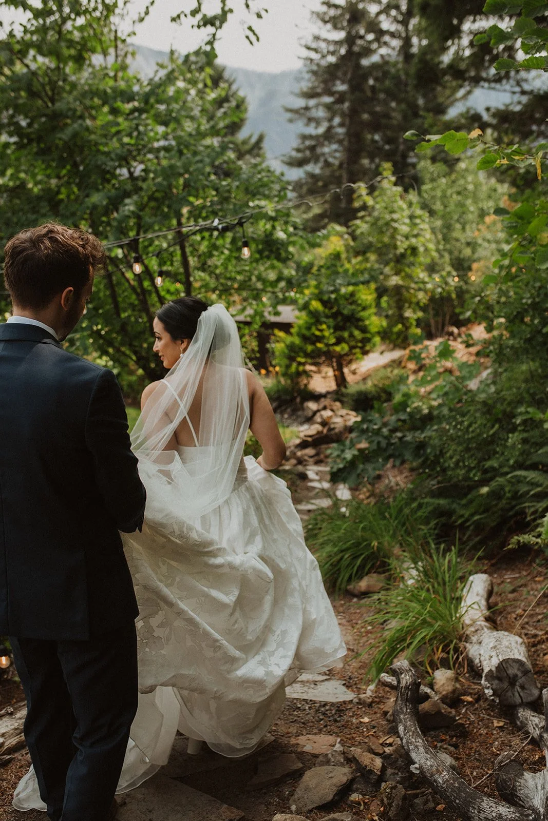 Groom smiles emotionally while holding the bride’s hands during their intimate first look on a green lawn surrounded by trees.