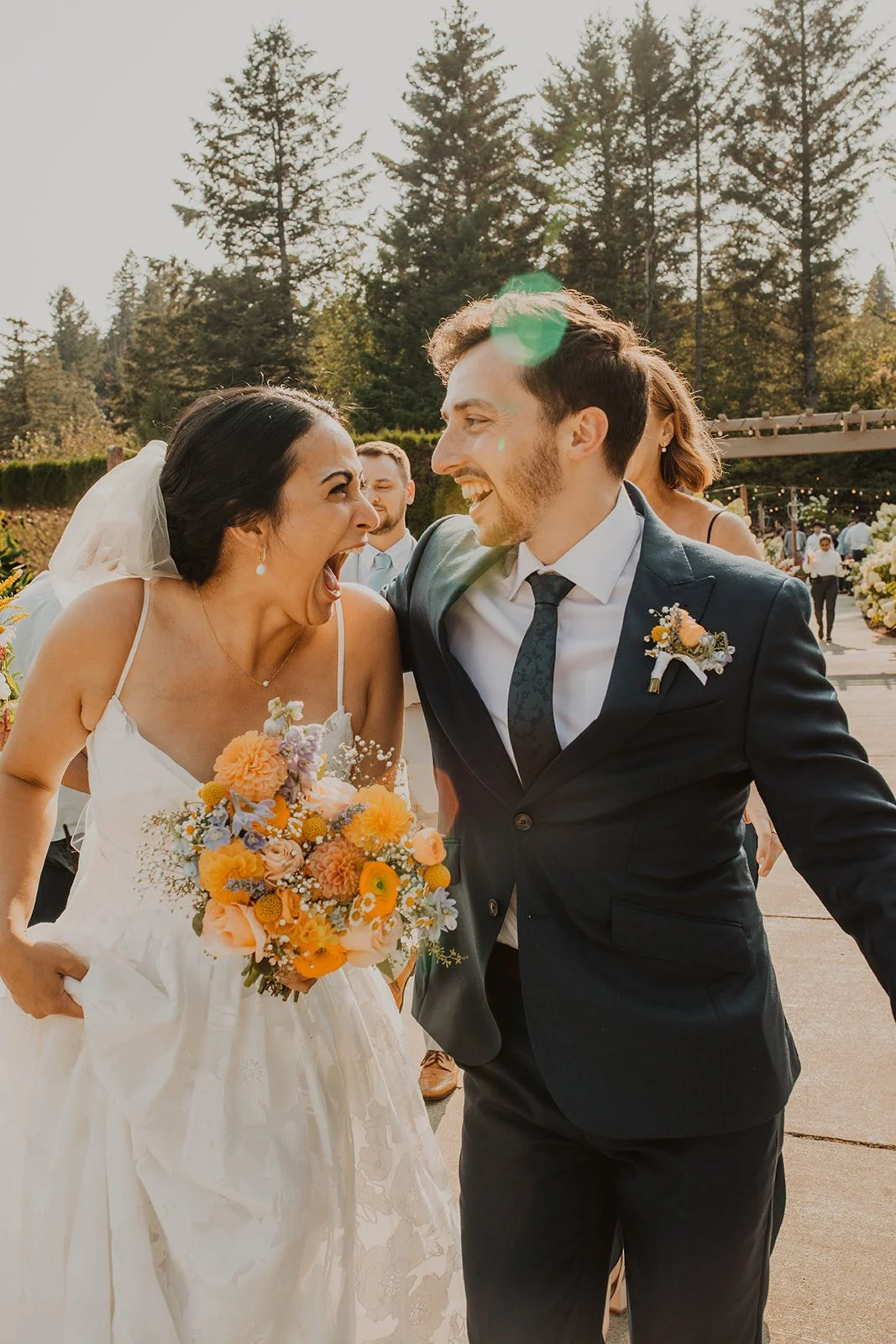 Joyful newlyweds laugh together while walking outdoors after their ceremony, with the bride holding a colorful bouquet in warm golden sunlight.