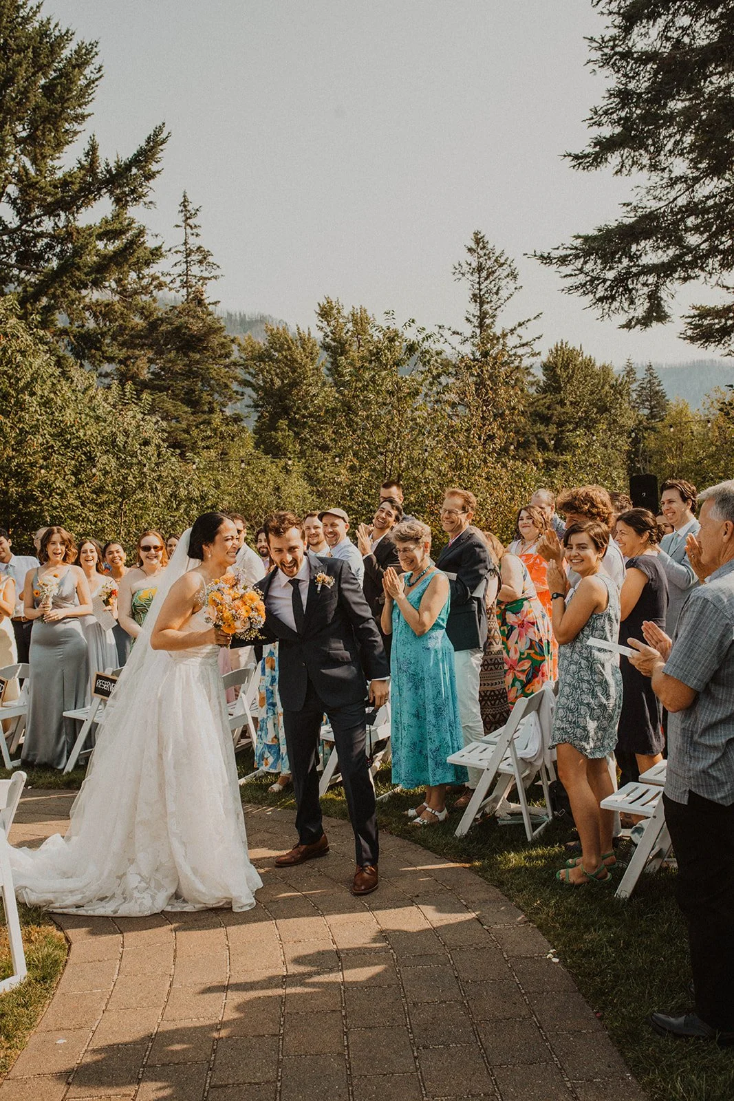 Newly married couple walks back down the aisle smiling and cheering as wedding guests stand and applaud during an outdoor garden ceremony.