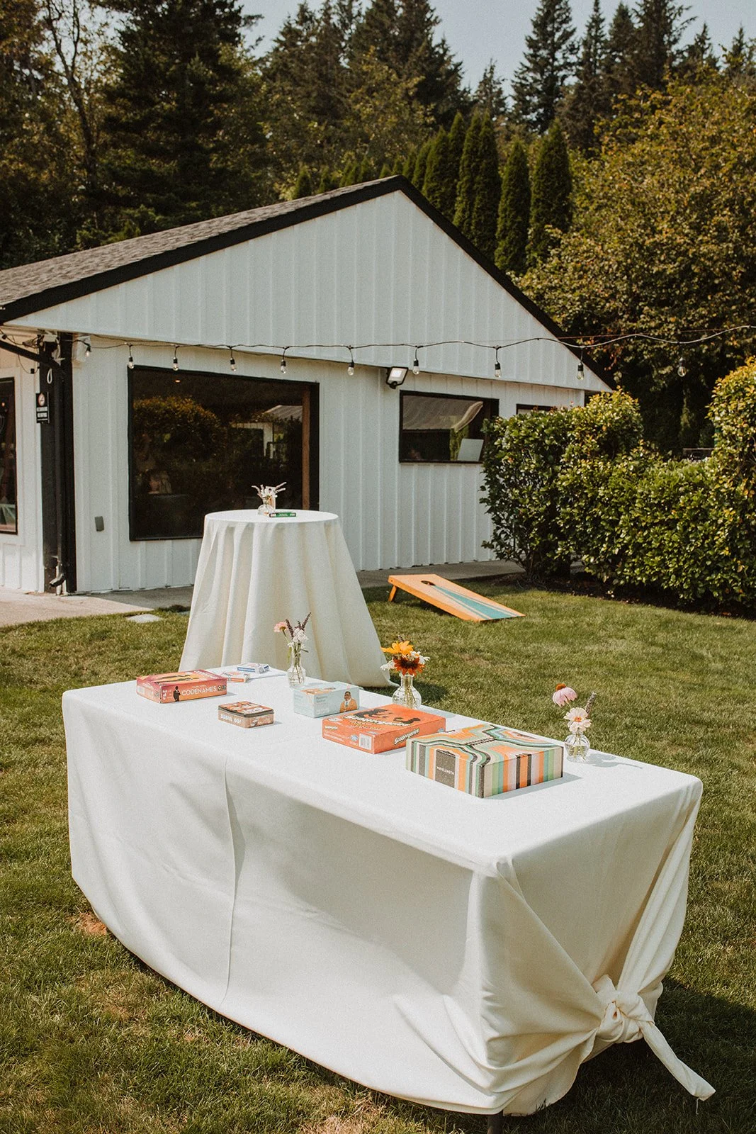 Outdoor wedding cocktail hour setup with lawn games, cocktail tables, and a white modern reception building surrounded by greenery and string lights