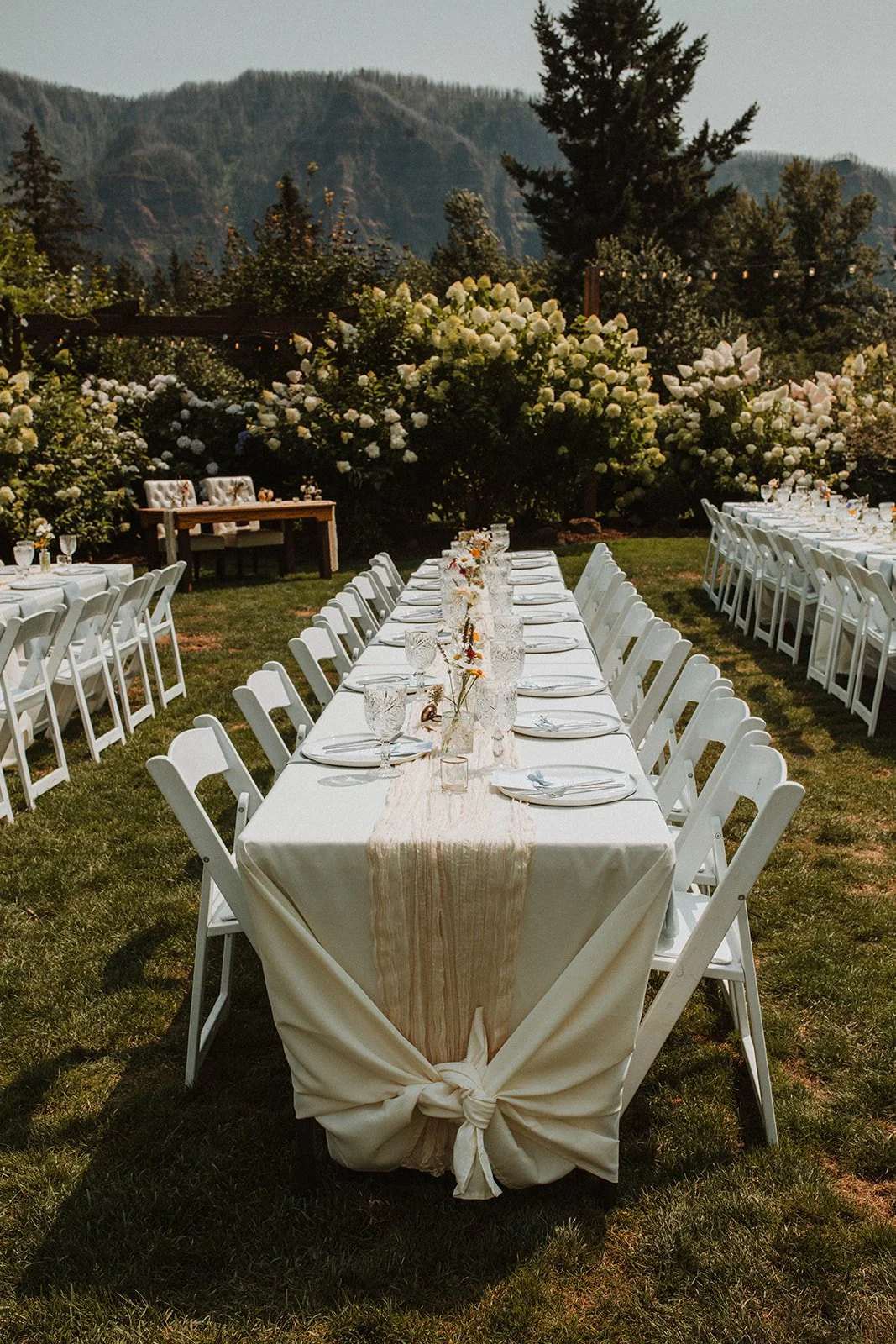 Elegant outdoor wedding reception tablescape with white folding chairs, neutral linens, glassware, and minimalist floral centerpieces set against mountain views and hydrangea gardens