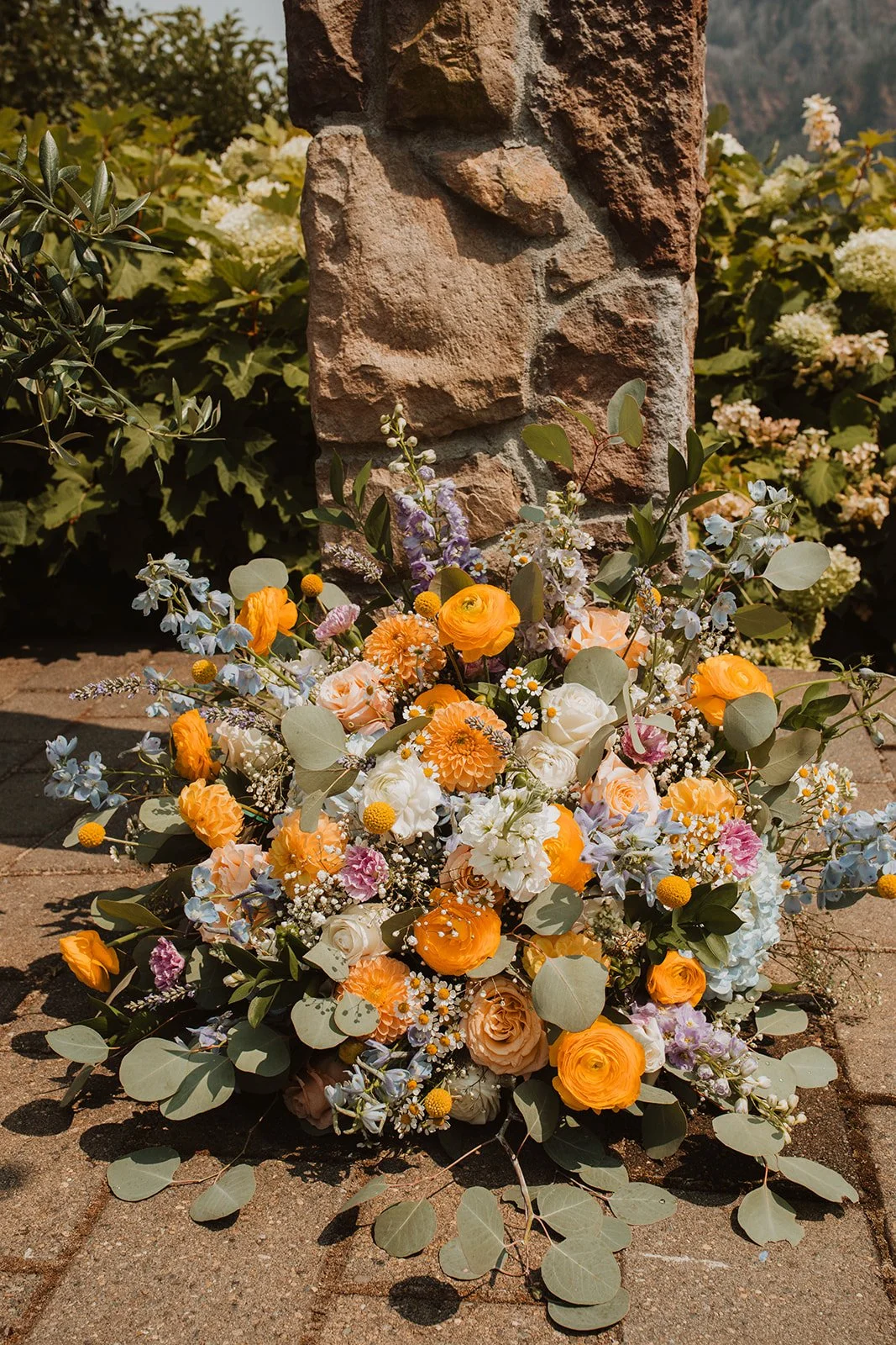 Colorful summer wedding ceremony floral arrangement featuring yellow ranunculus, peach roses, blue delphinium, chamomile, eucalyptus, and pastel blooms displayed beside a stone pillar outdoors