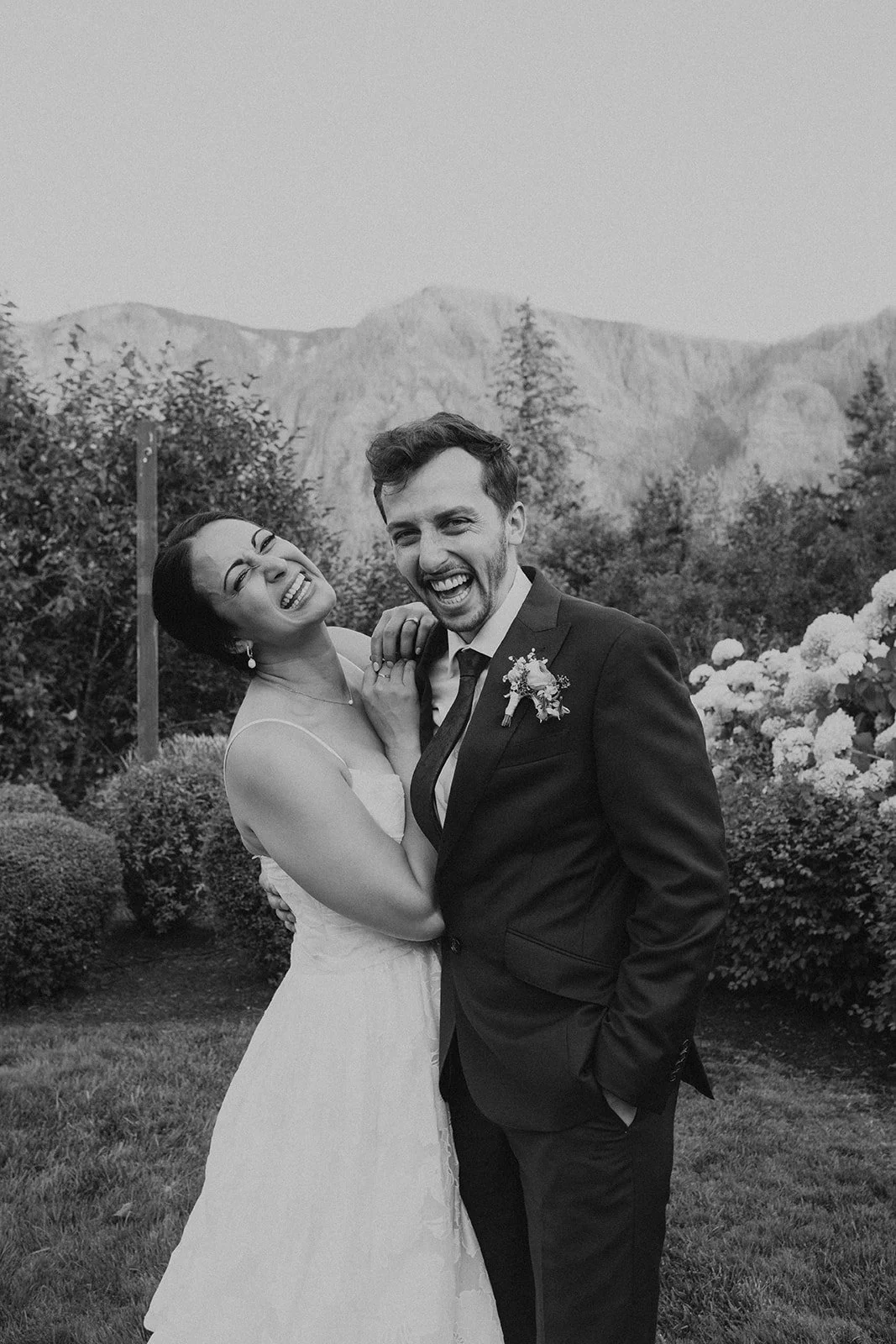 Black-and-white portrait of a joyful bride and groom laughing together outdoors with mountains and hydrangeas in the background.