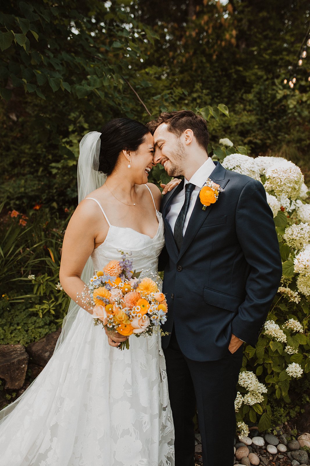 Bride and groom smiling forehead to forehead while holding a colorful summer bouquet beside blooming white hydrangeas at an outdoor wedding venue.