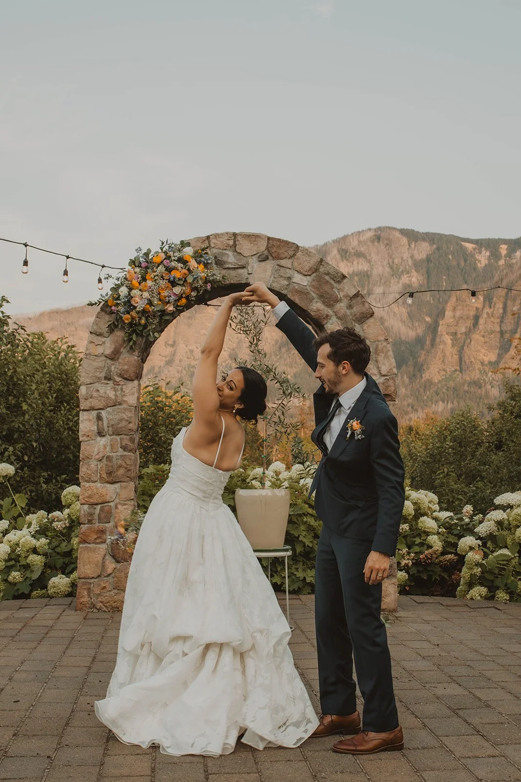 Bride twirls under a stone floral arch while dancing with the groom during sunset portraits at a mountain garden wedding venue.