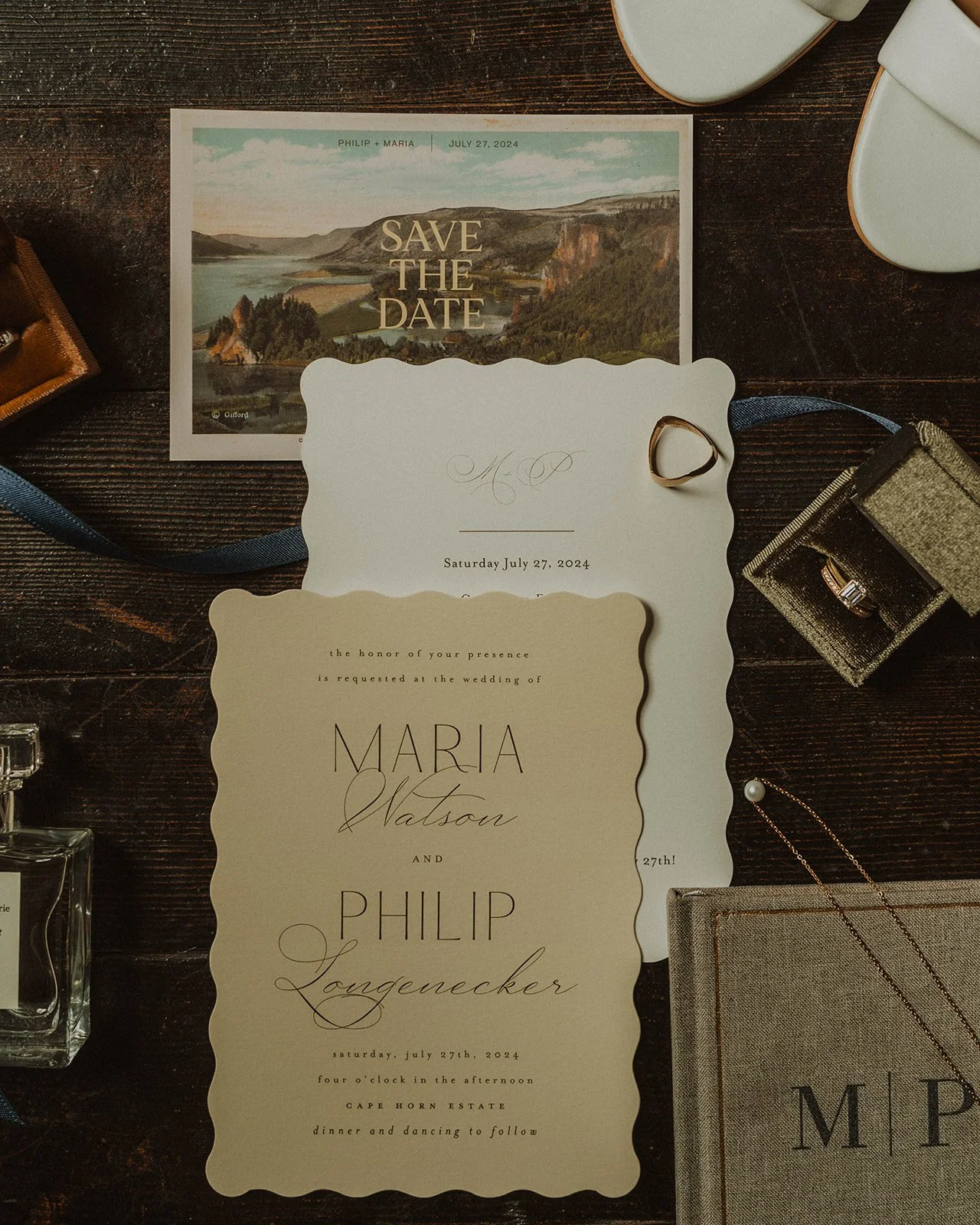 Elegant wedding invitation suite with scalloped-edge stationery, save the date card, wedding rings, and perfume styled on a dark wooden table.
