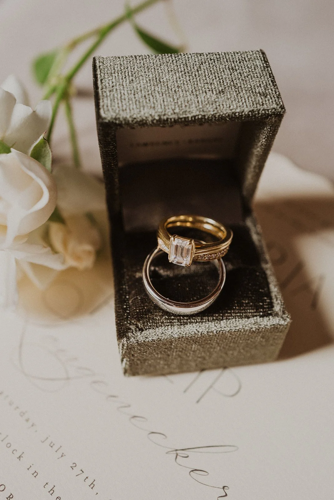 Emerald-cut engagement ring and wedding band displayed inside a textured black ring box on top of wedding invitation stationery.