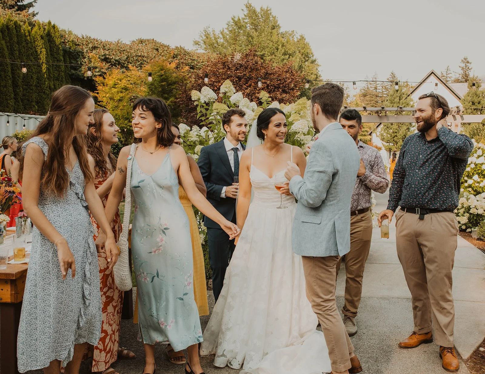 Bride greeting and hugging wedding guests during cocktail hour at an outdoor reception with warm golden sunlight and string lights overhead.