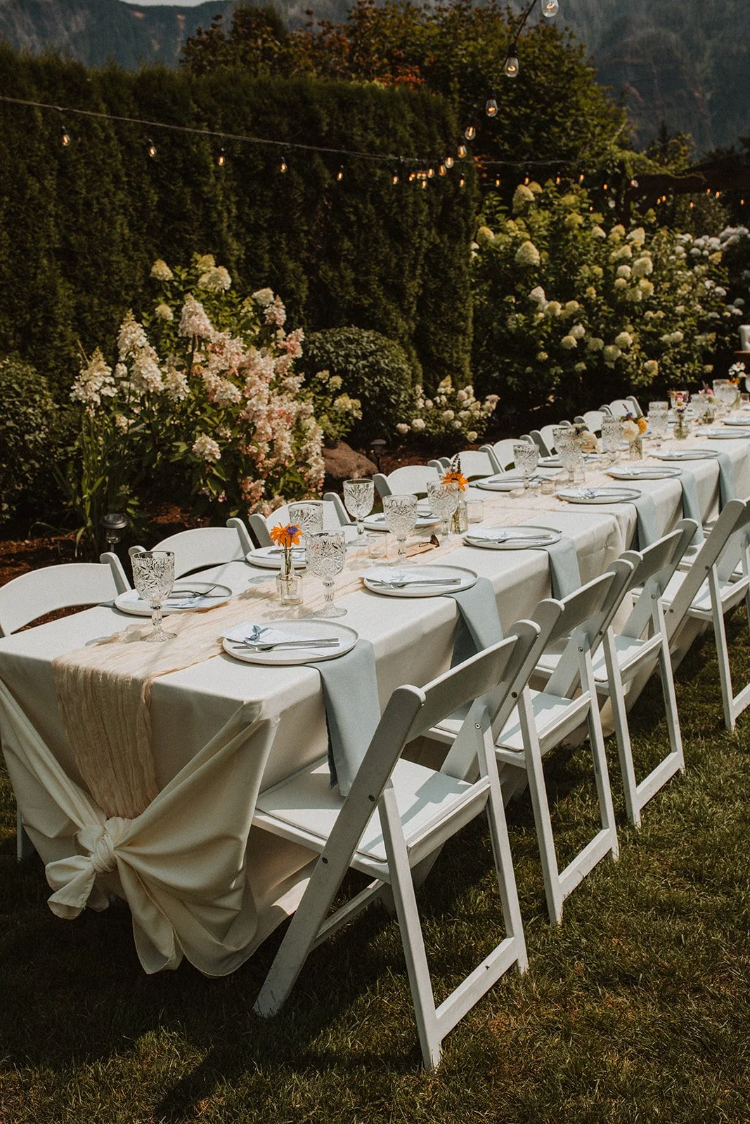 Long outdoor wedding reception table with white folding chairs, soft neutral linens, crystal glassware, and string lights surrounded by hydrangea gardens.