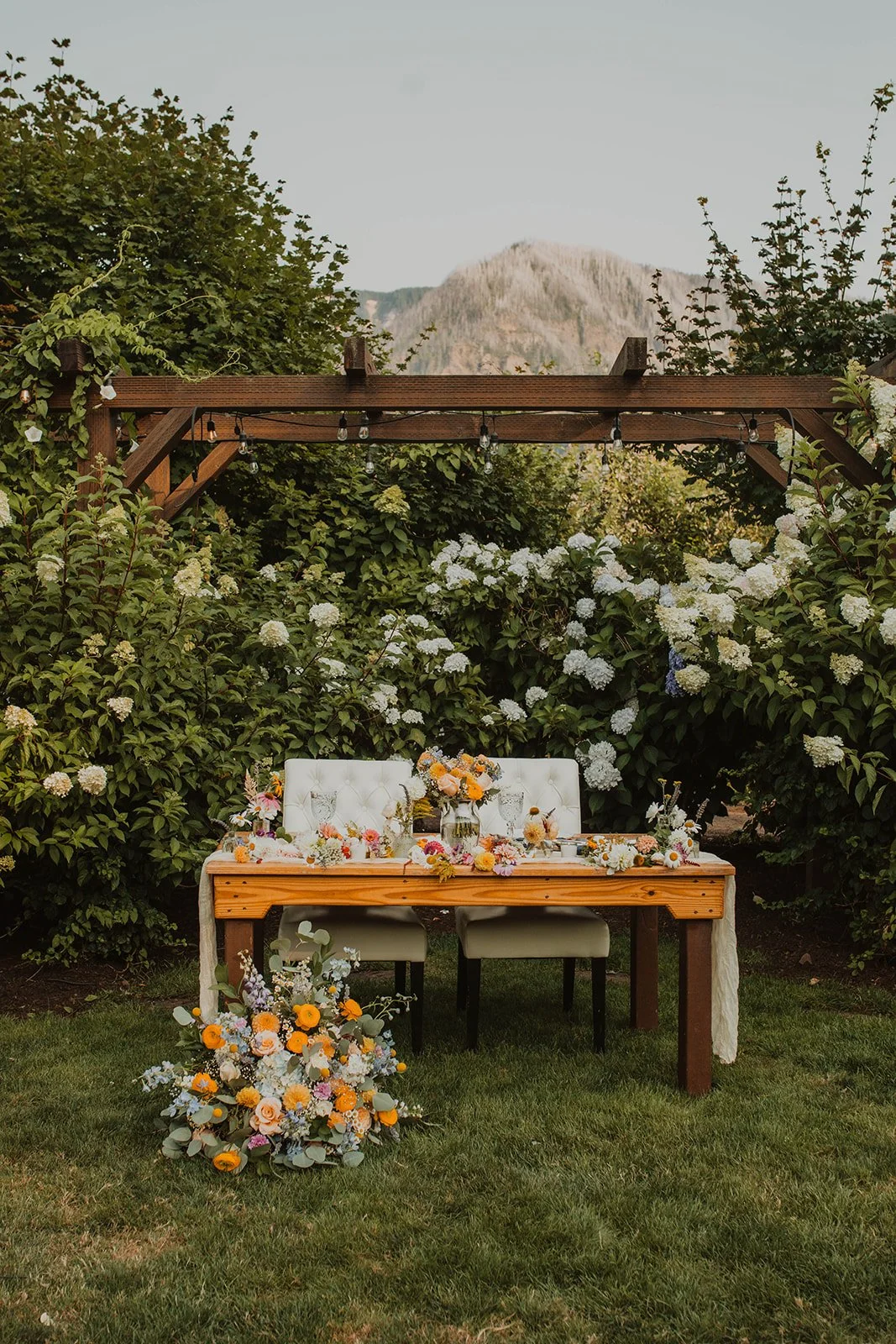 Elegant outdoor sweetheart table decorated with colorful florals beneath a wooden pergola surrounded by hydrangeas and mountain views.
