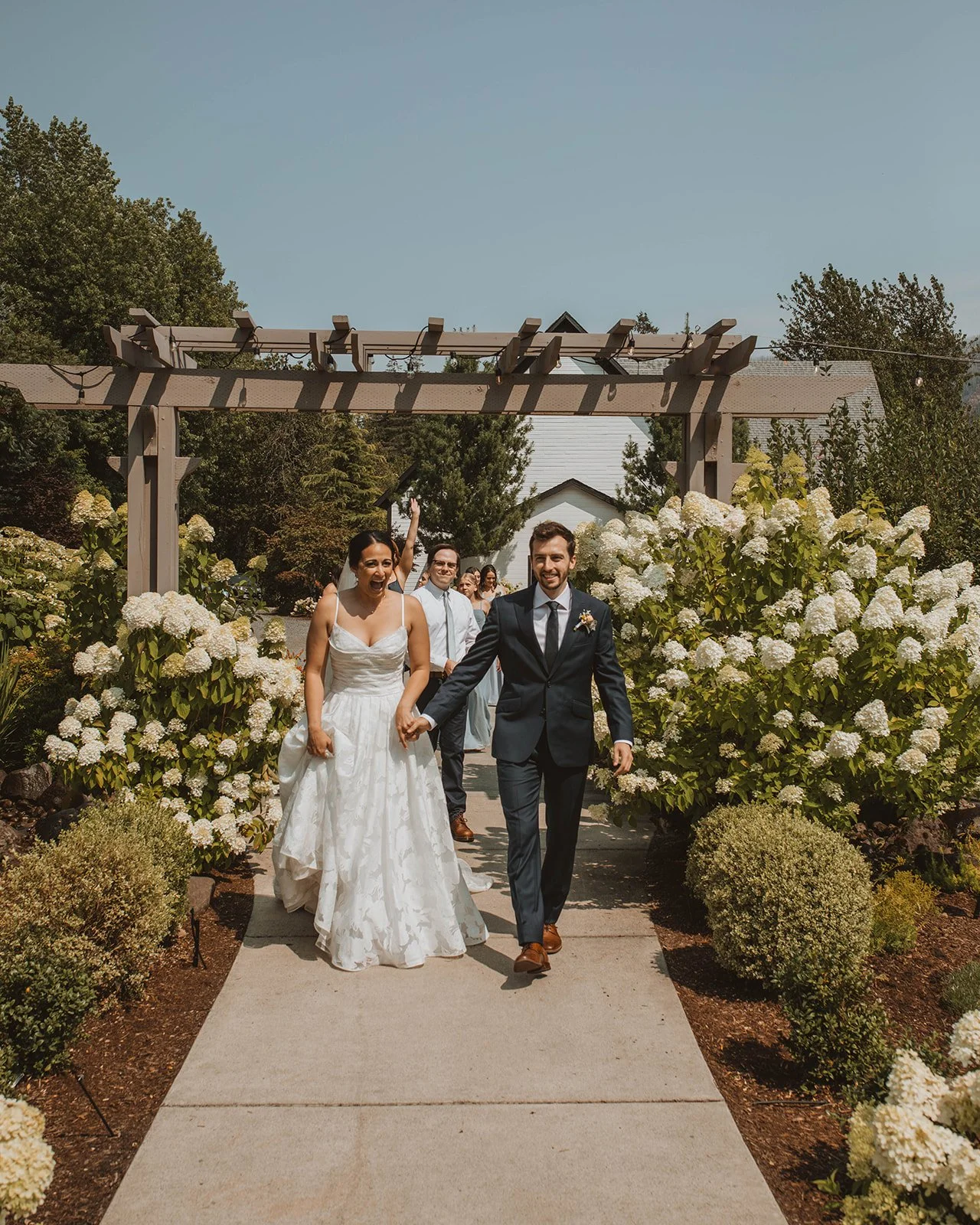 Joyful newlywed couple walking hand in hand through a garden pathway lined with white hydrangeas and pergolas at an outdoor wedding venue.