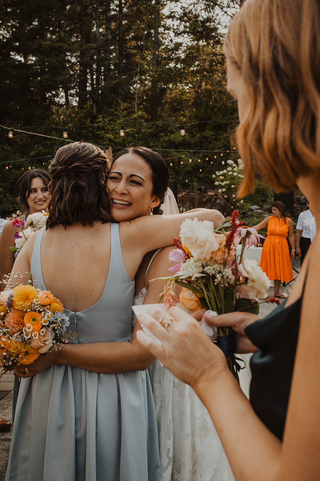Emotional moment as the bride hugs a bridesmaid holding a colorful bouquet during an outdoor wedding reception surrounded by guests and string lights.