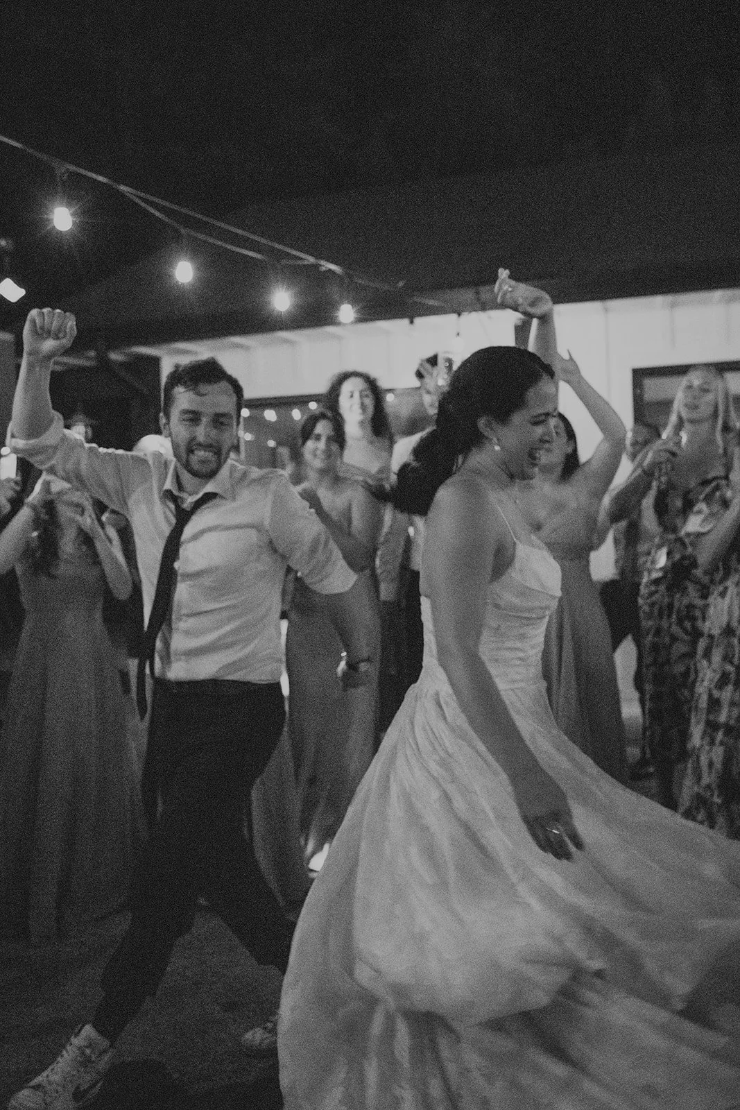 Black-and-white candid photo of the bride and groom dancing energetically on the reception dance floor surrounded by cheering wedding guests under string lights.