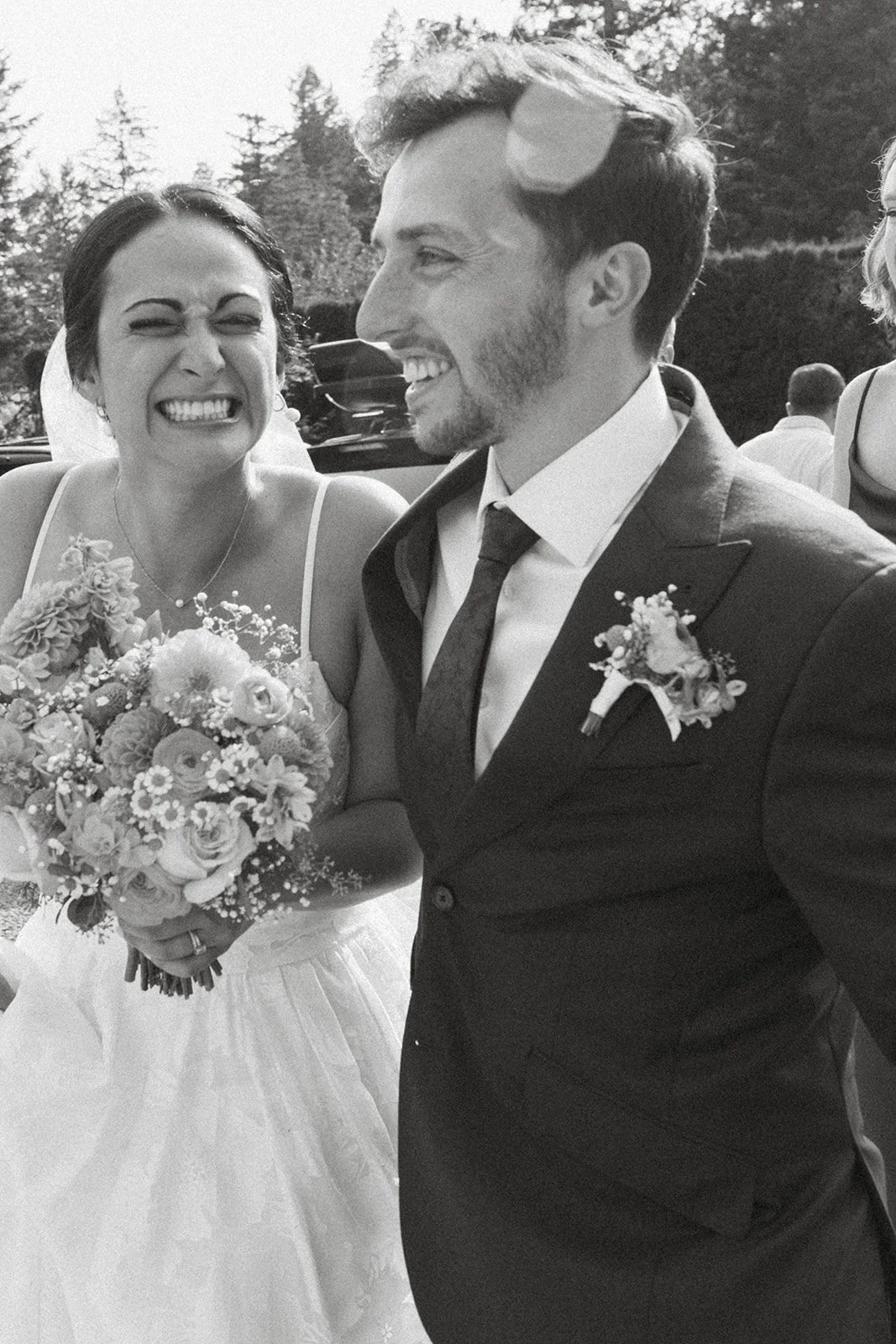 Black-and-white candid portrait of the newlywed couple smiling and laughing together while holding a colorful wedding bouquet outdoors.