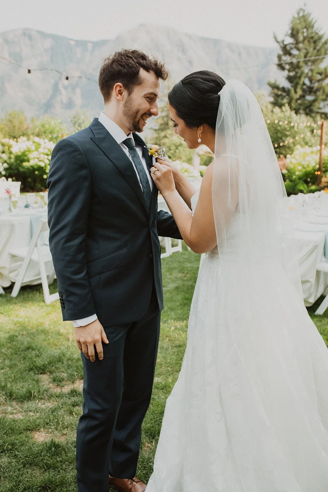 Bride adjusting the groom’s boutonniere while smiling together outdoors before their wedding reception with mountain views in the background.