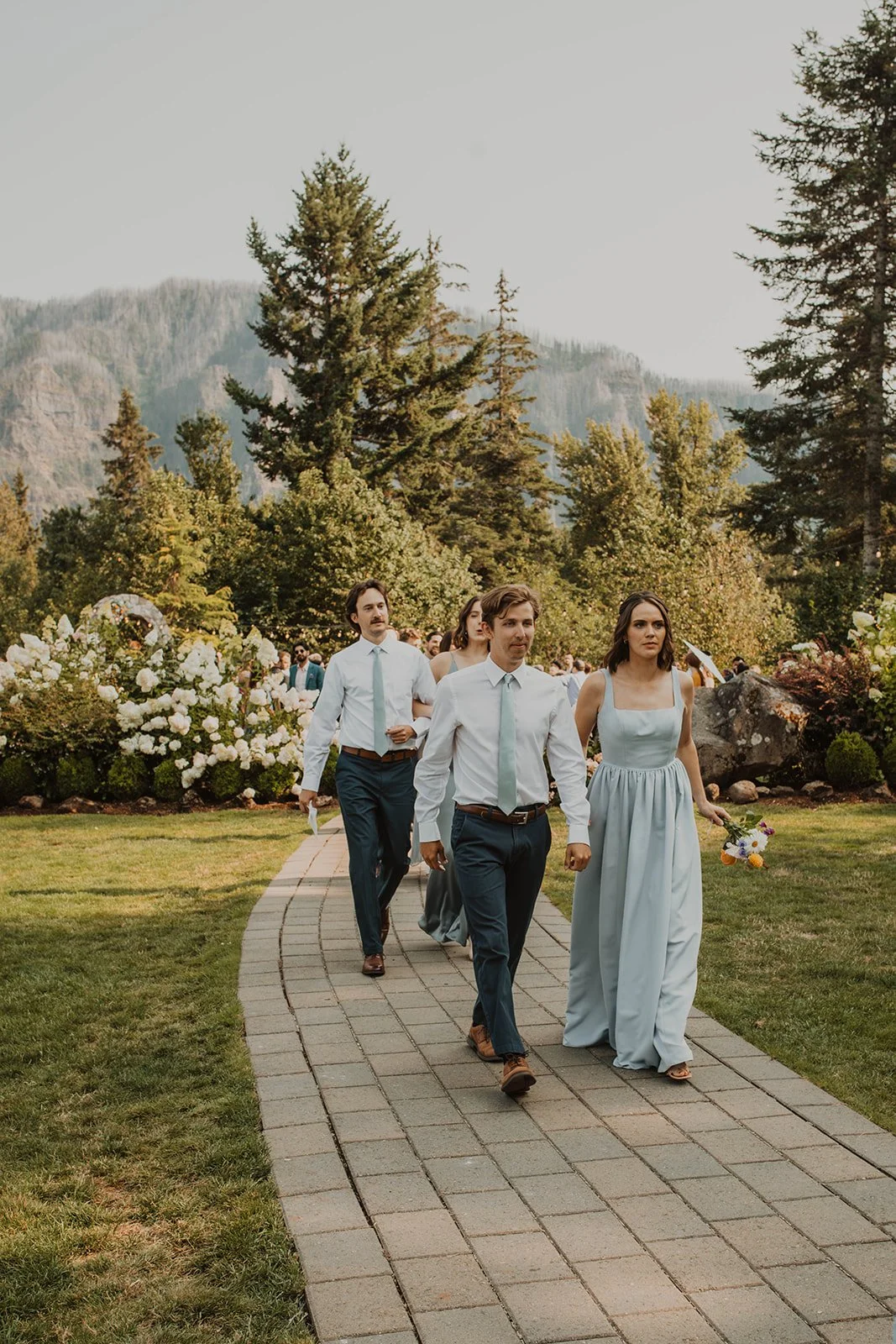 Bridesmaids and groomsmen walking down a curved garden pathway at an outdoor mountain wedding venue before the ceremony.