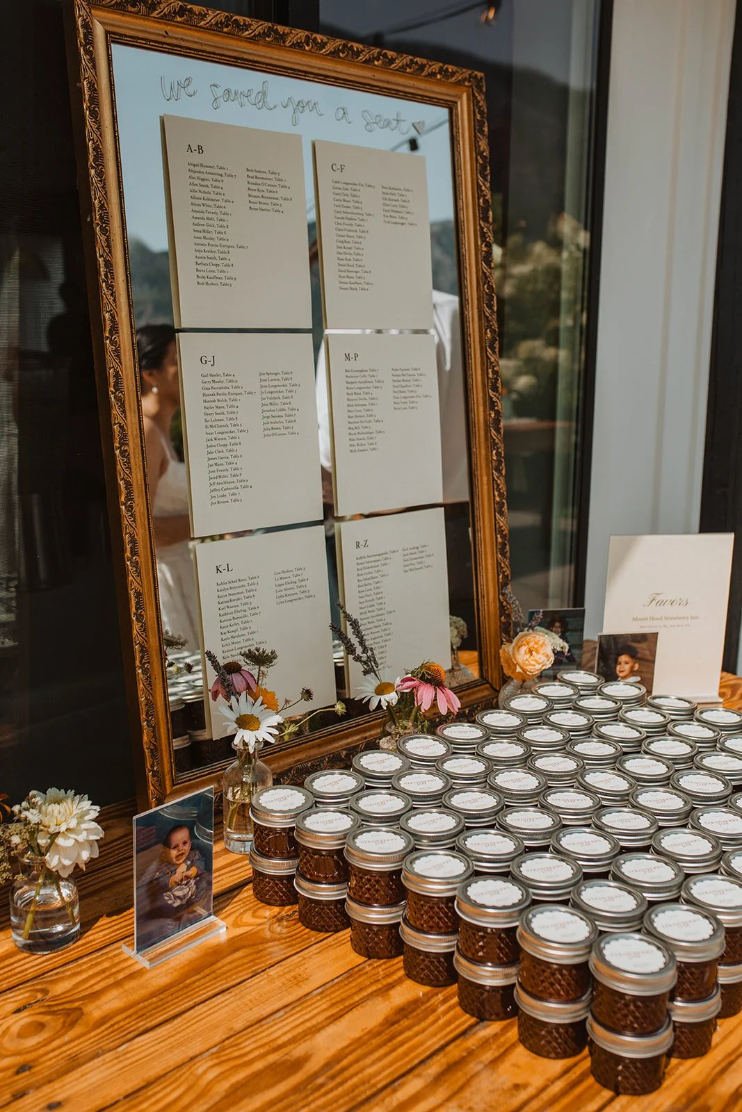 Framed wedding seating chart displayed beside mini jam jar wedding favors and floral bud vases on a rustic wooden table.