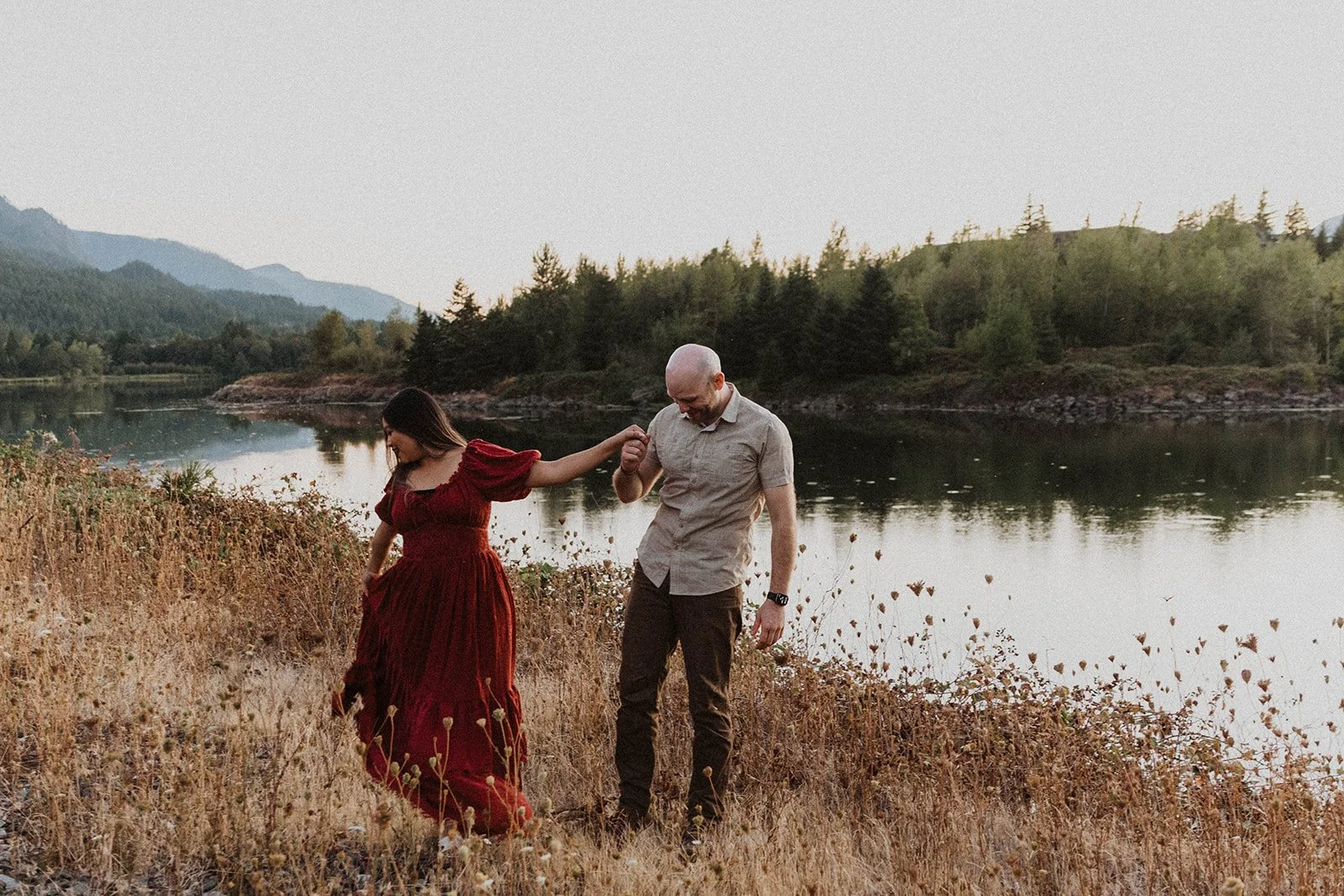 Engaged couple walking hand in hand along a lakeshore surrounded by mountains during an outdoor engagement session at sunset.
