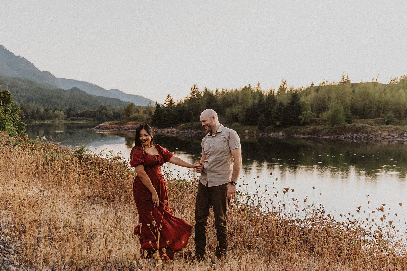 Playful engagement portrait beside a river with forested mountains creating a scenic Mt Hood backdrop