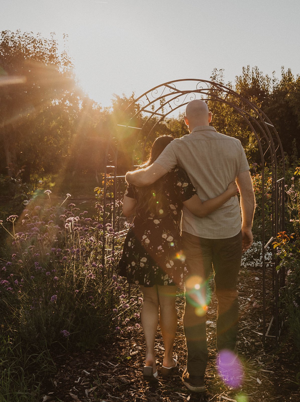 Couple walking arm in arm through a garden archway at sunset during a romantic engagement session.