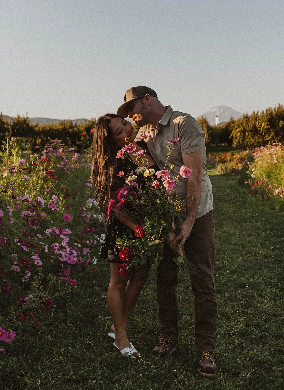 Engaged couple standing together in a flower field holding a freshly gathered bouquet during sunset.