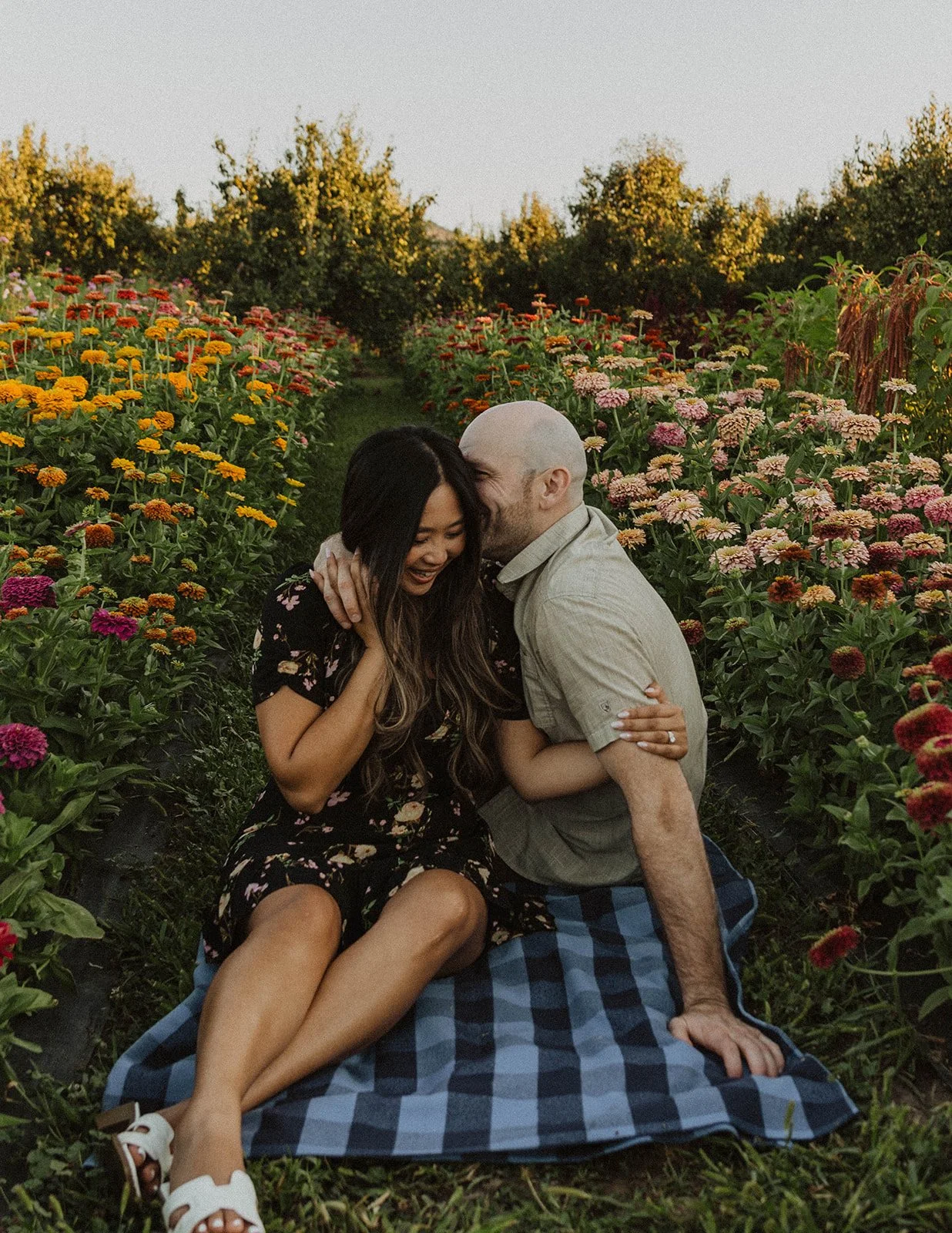 Couple laughing together on a picnic blanket between rows of vibrant flowers during an engagement session.