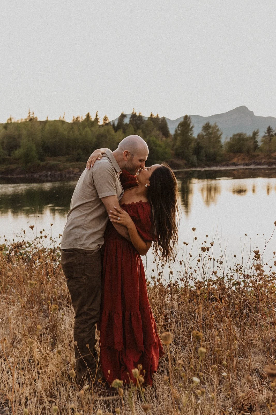 Couple sharing a kiss beside a riverside meadow during golden hour with mountain views near Mt Hood.