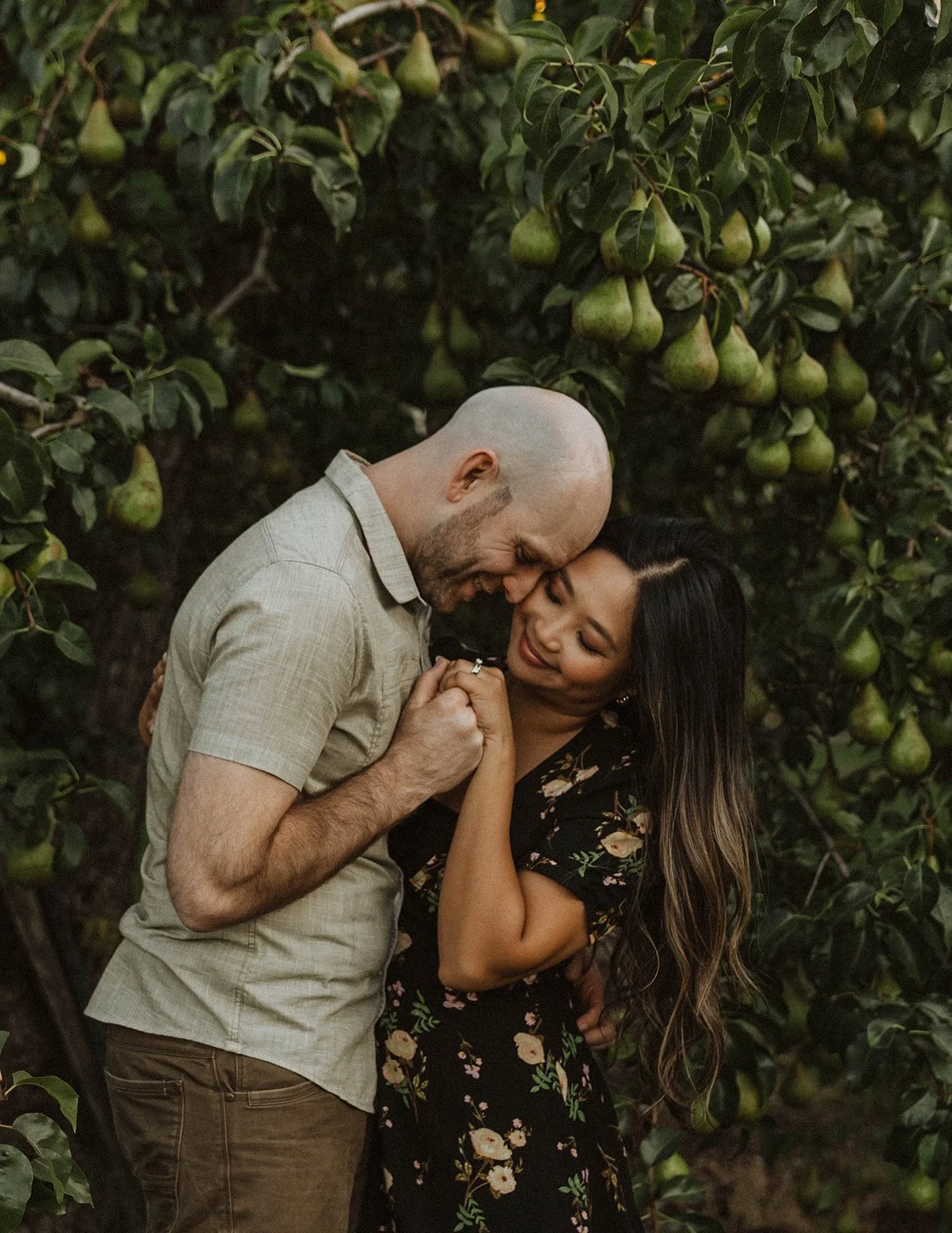 Engaged couple sharing a quiet moment beneath fruit-filled pear trees during a romantic orchard engagement session.