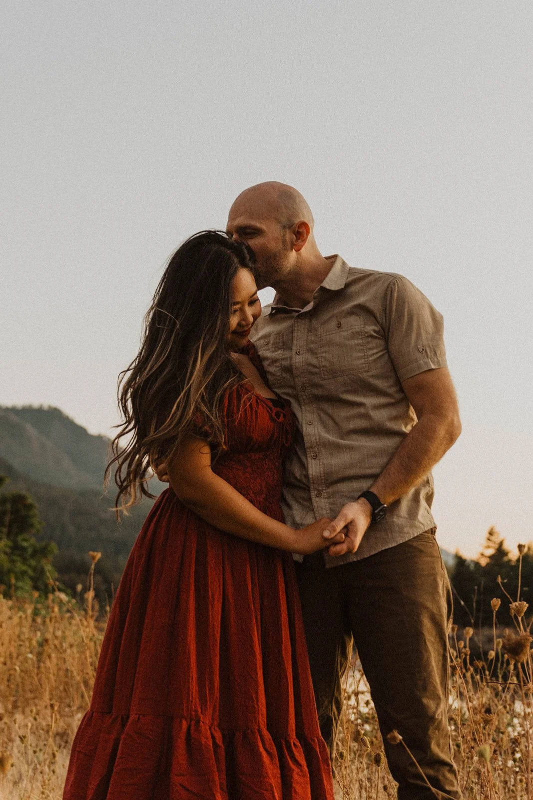 Groom kissing his partner's forehead beside a mountain lake during a golden hour engagement session.