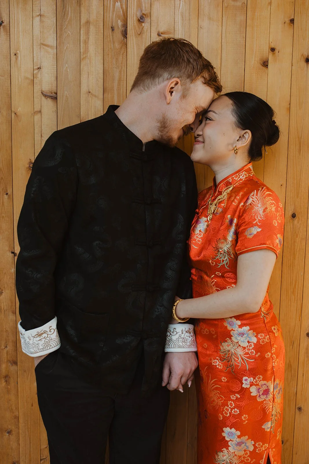 Bride and groom share a quiet moment in traditional Chinese wedding attire against a warm wooden backdrop.