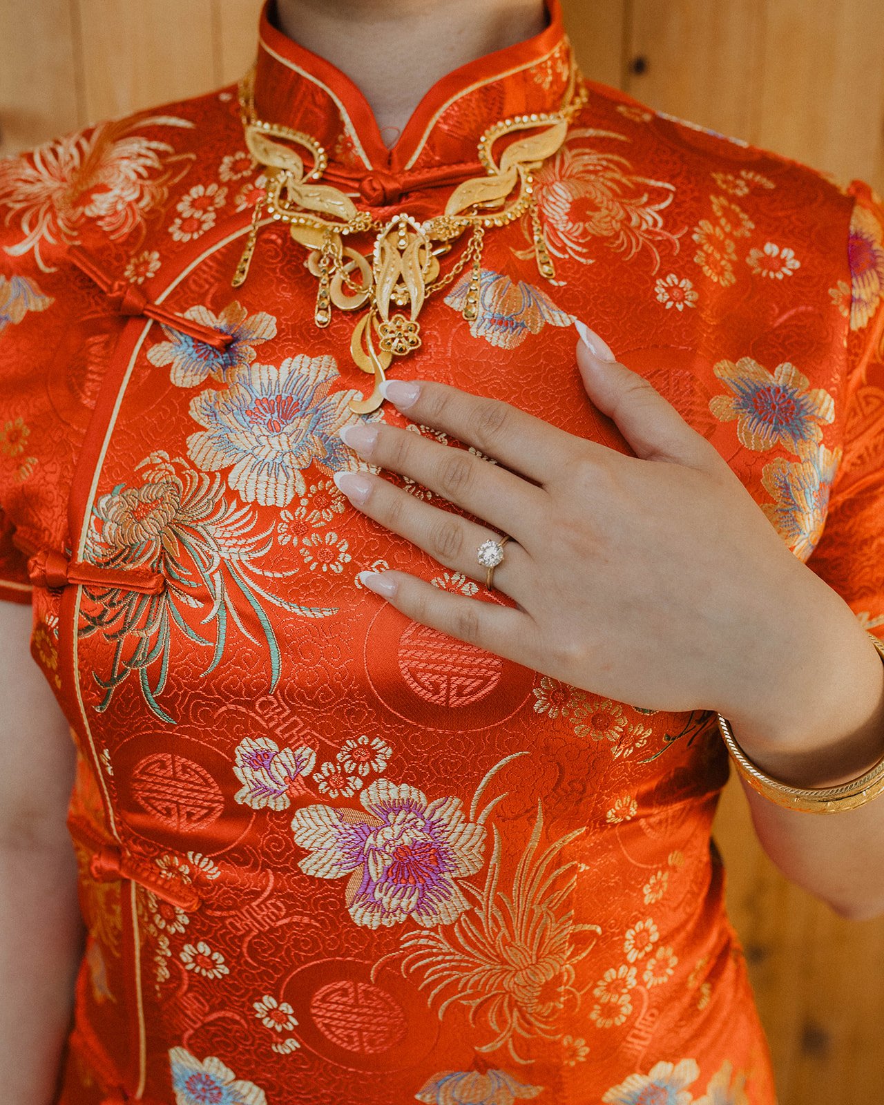 Close-up of bride’s engagement ring and gold jewelry against a red embroidered cheongsam during a traditional Chinese wedding celebration.