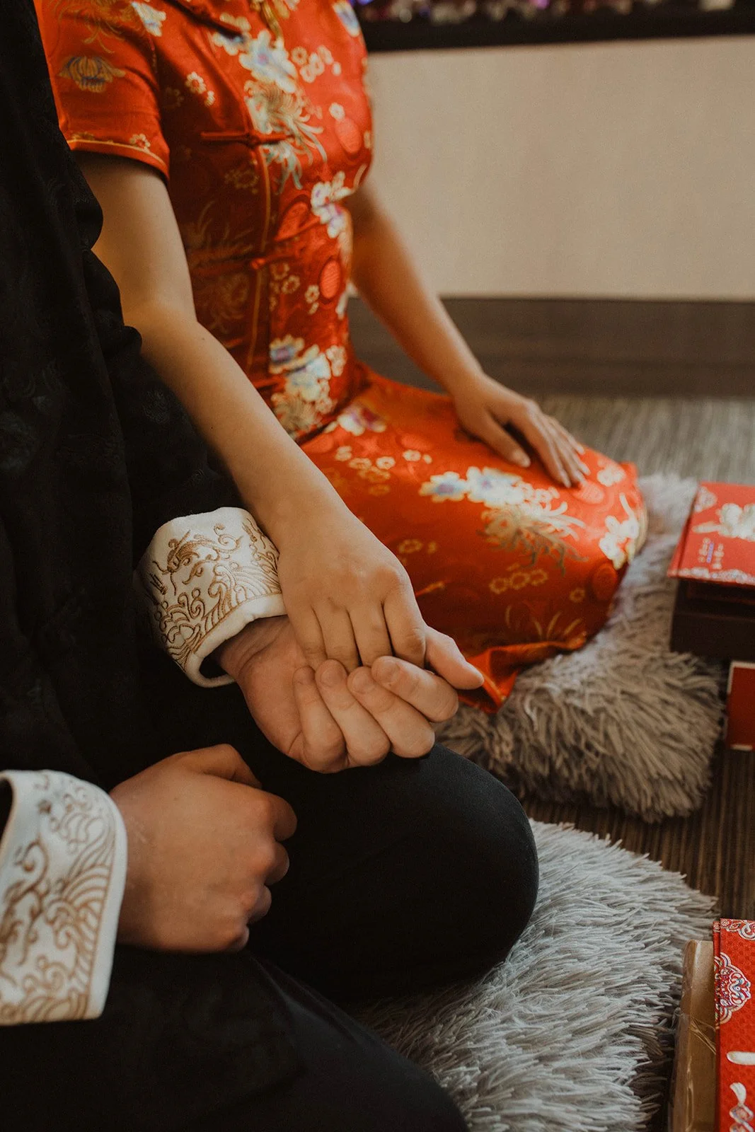 Close-up of bride and groom holding hands during a traditional Chinese tea ceremony while wearing red and black ceremonial attire.