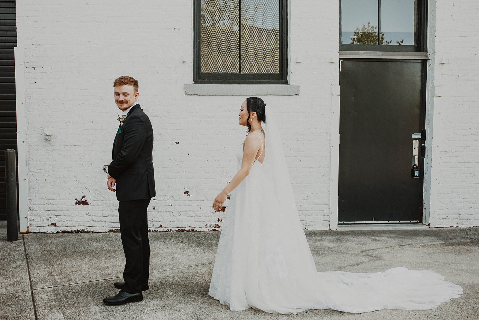 Bride approaches her groom from behind before their first look outside an industrial wedding venue with white brick walls.
