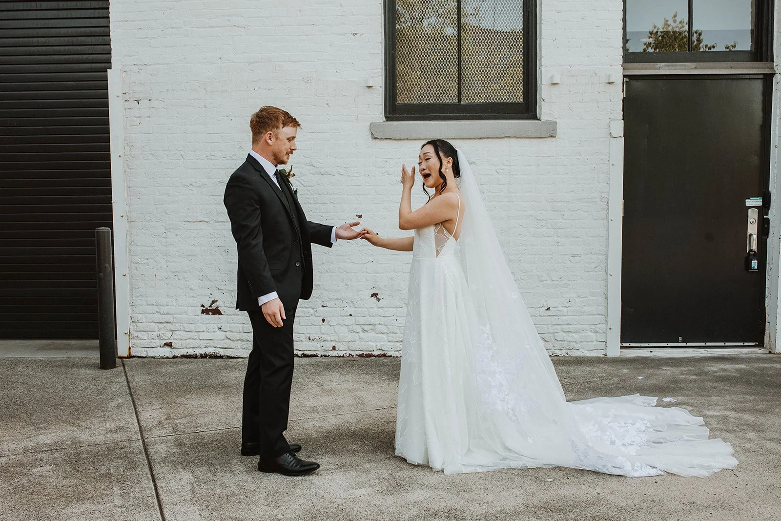 Bride reacts emotionally during a first look as she reaches for her groom’s hand outside their wedding venue.