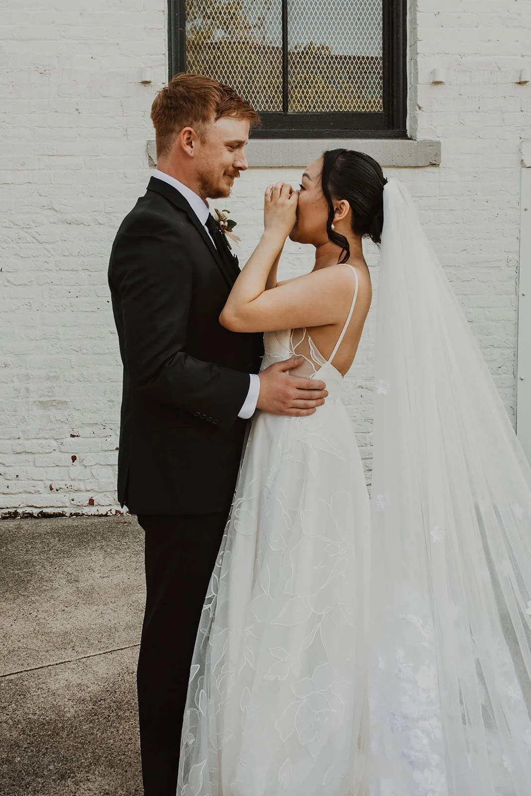 Bride wipes away tears while standing face-to-face with her groom during an emotional wedding portrait outside an industrial wedding venue.