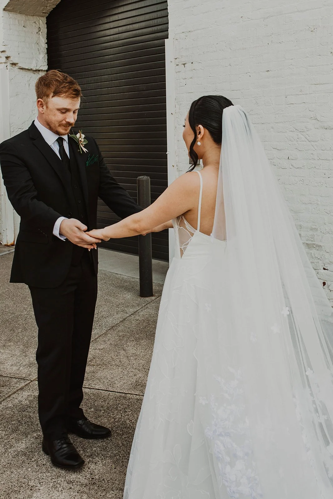 Bride and groom hold hands and smile at one another during wedding portraits outside an industrial-style wedding venue.