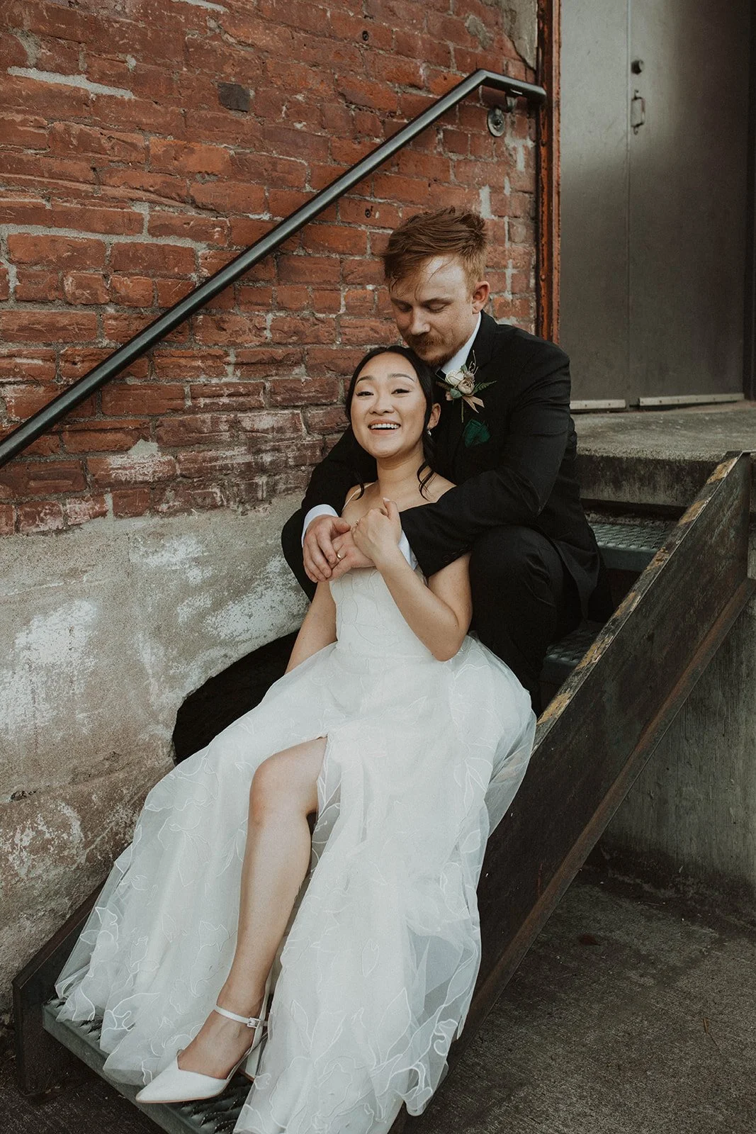 Bride and groom sit together on an industrial metal staircase, sharing a quiet moment during wedding portraits at an urban venue.