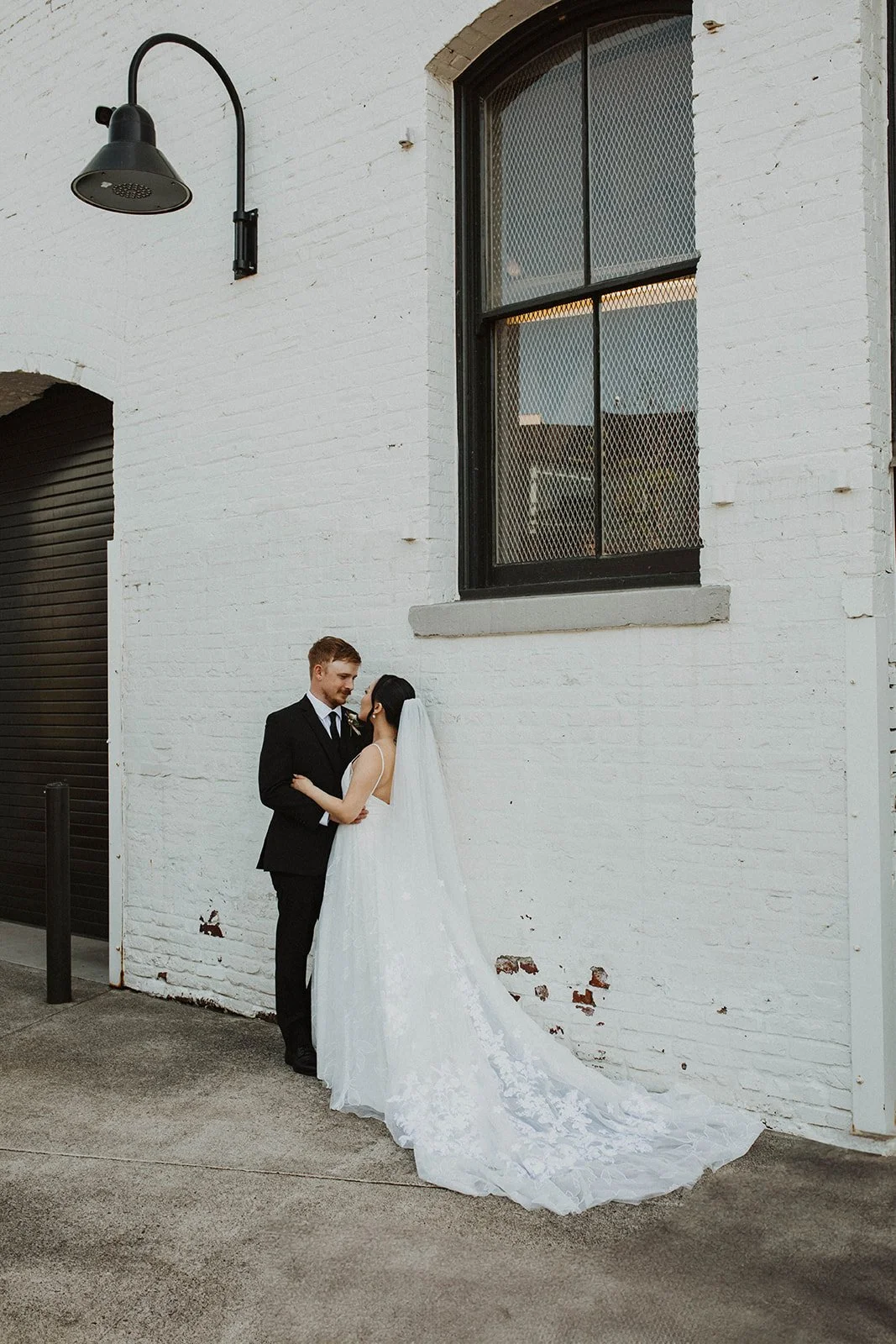 Bride and groom embrace during wedding portraits against a white brick wall outside an industrial wedding venue.