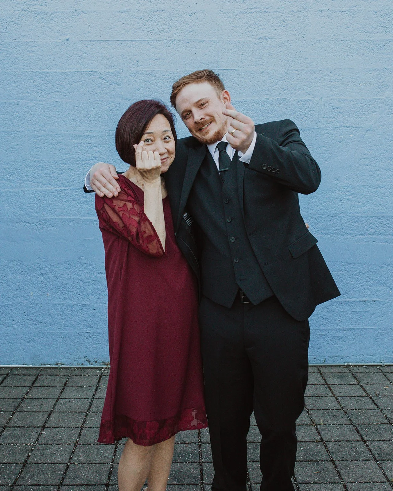Groom and his mother share a playful moment during wedding portraits, making finger heart gestures while standing against a blue wall outside a Portland wedding venue.